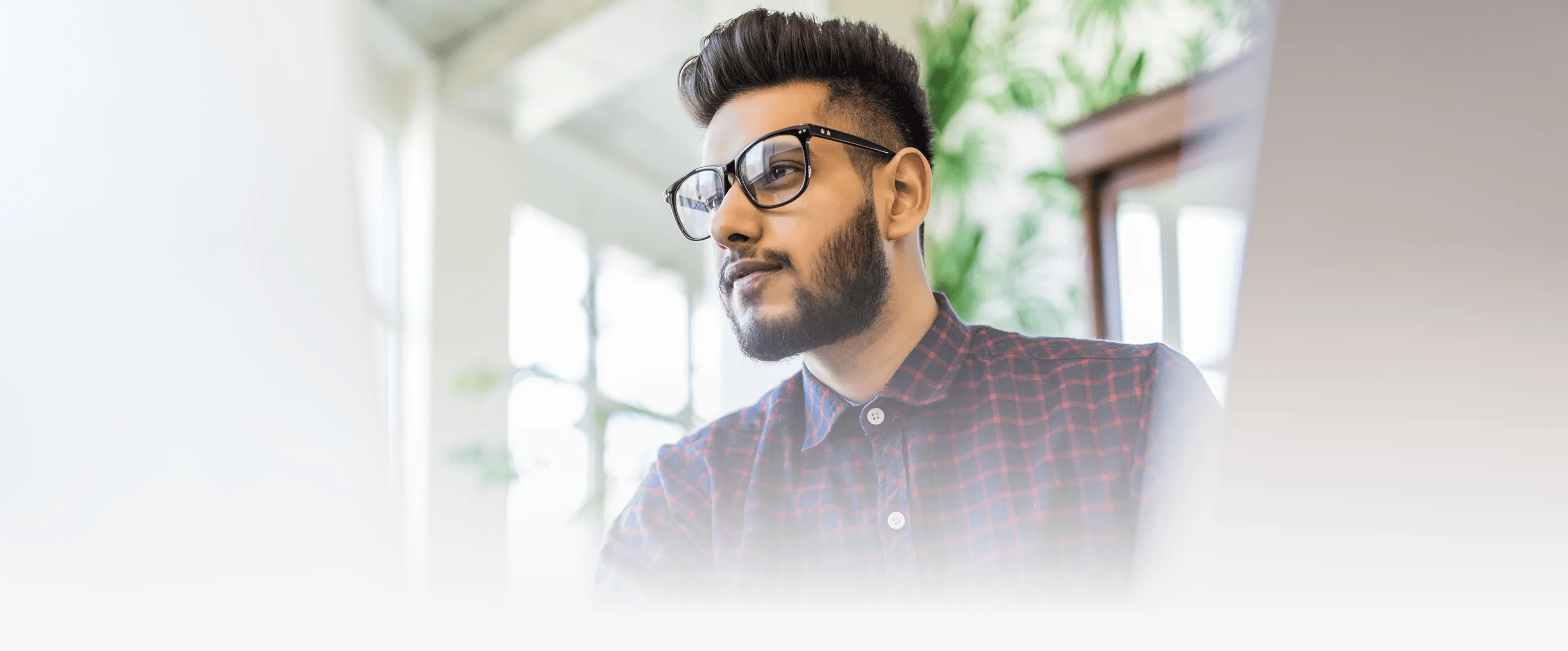 A man in a checkered shirt sitting at a desk, focused and wearing glasses while working at a computer.