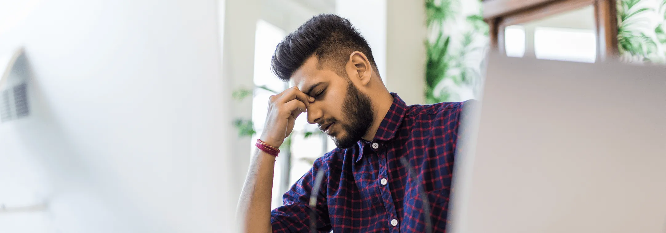 A man in a checkered shirt sitting at a desk, holding his forehead in frustration.