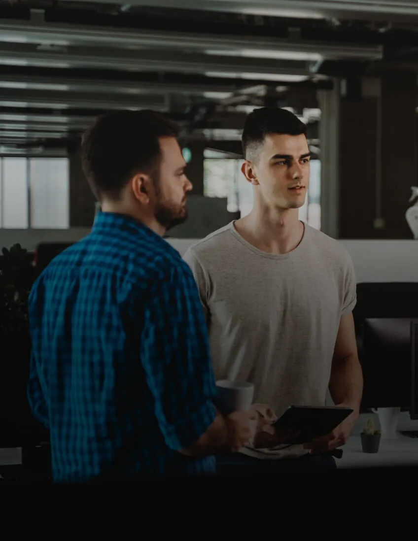 Two men in an office having a discussion, one holding a tablet and the other holding a cup.