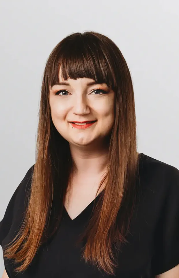 Smiling woman with long straight brown hair and bangs wearing a black V-neck shirt against a plain light background.