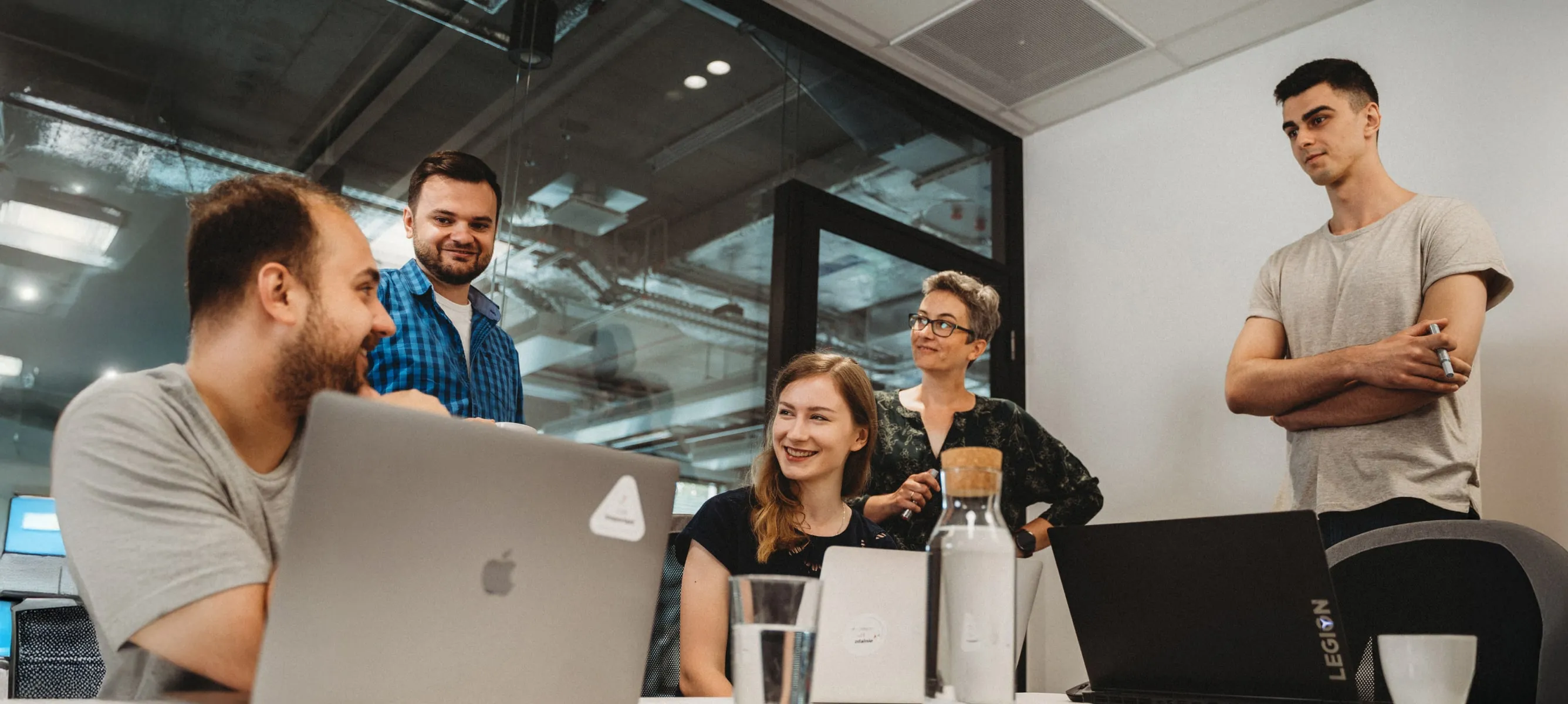 Five colleagues working together in a modern office, two sitting with laptops and three standing, engaged in a discussion.