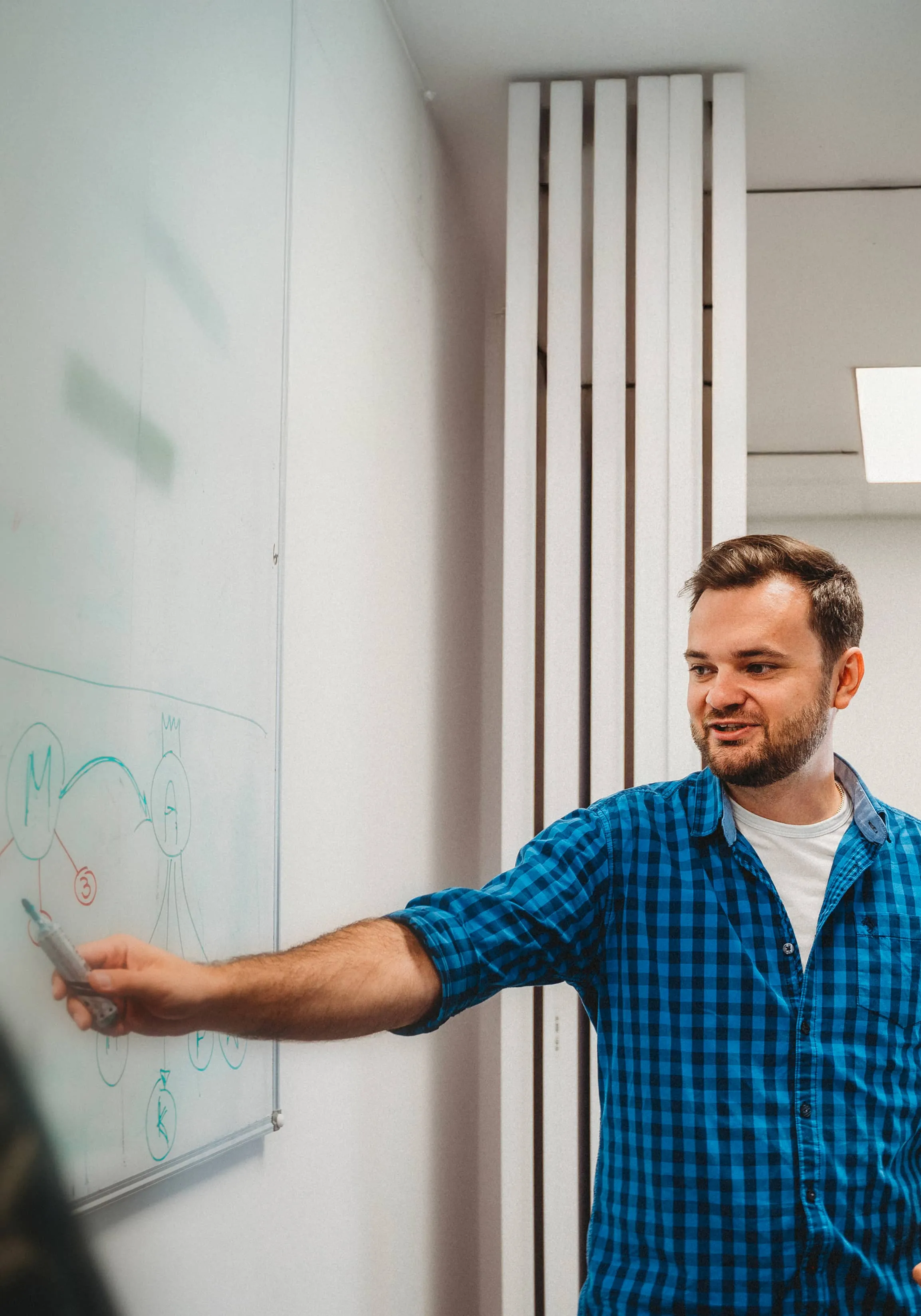 Man in a blue checkered shirt explaining a diagram on a whiteboard with a marker in hand.