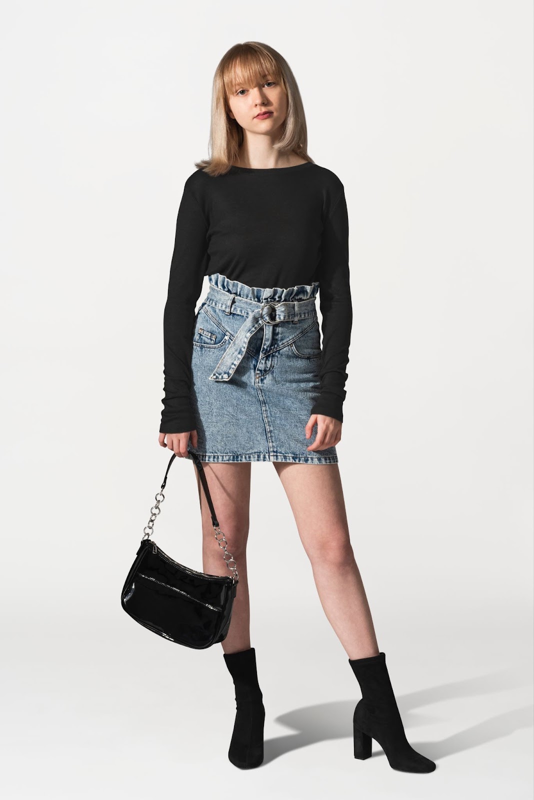 A woman wearing a high-waisted denim mini skirt with a black long-sleeve t-shirt, boots, and bag.