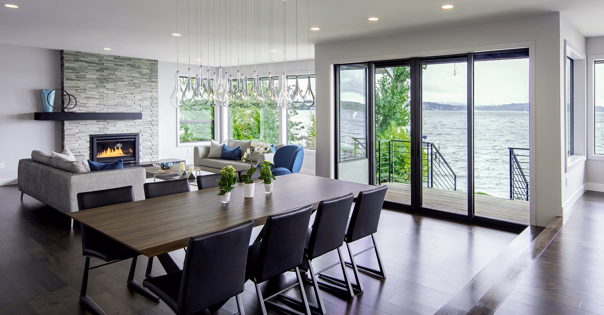 A modern open-concept living and dining area with dark wood floors and light gray walls. The living room features a gray sofa with blue accent pillows and a blue swivel chair, centered around a gas fireplace with a textured stone wall. The dining area has a dark wood table with black chairs and a contemporary chandelier with glass pendants. Large sliding glass doors and windows provide a scenic view of a lake, with a deck and railing leading to the water. The room is bright with natural light and recessed ceiling lights, creating a spacious and airy atmosphere.