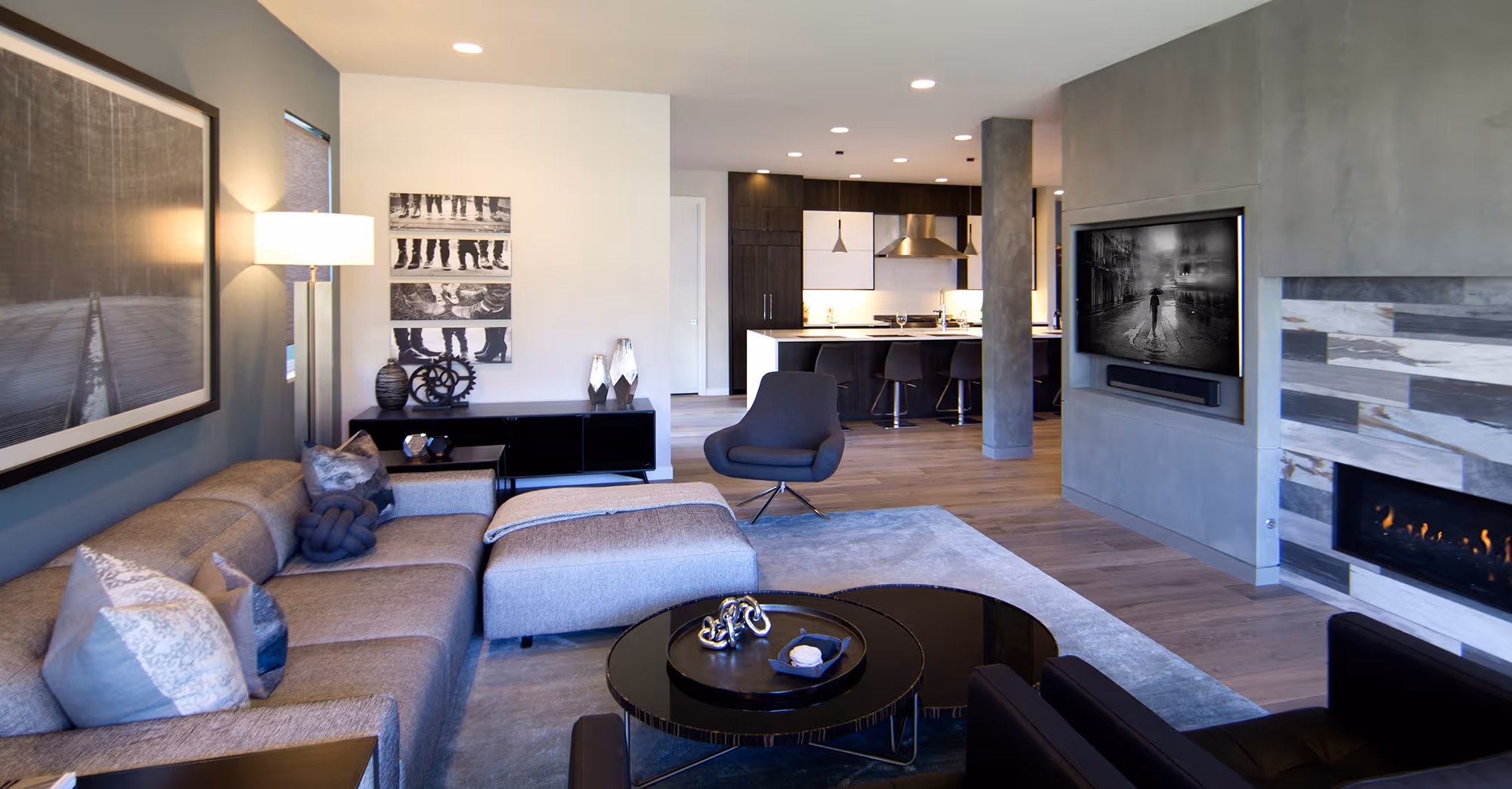 A contemporary open-concept living and dining area with dark wood floors and light gray walls. The living room features a cozy gray sofa, blue accent pillows, and a blue swivel chair, with a modern gas fireplace set in a textured stone wall. The dining area has a dark wood table with black chairs and a modern chandelier with glass pendant lights. Large sliding glass doors and windows provide an expansive view of a lake, and a deck with a railing is visible outside. The room is bright and airy, with ample natural light and recessed ceiling lights.