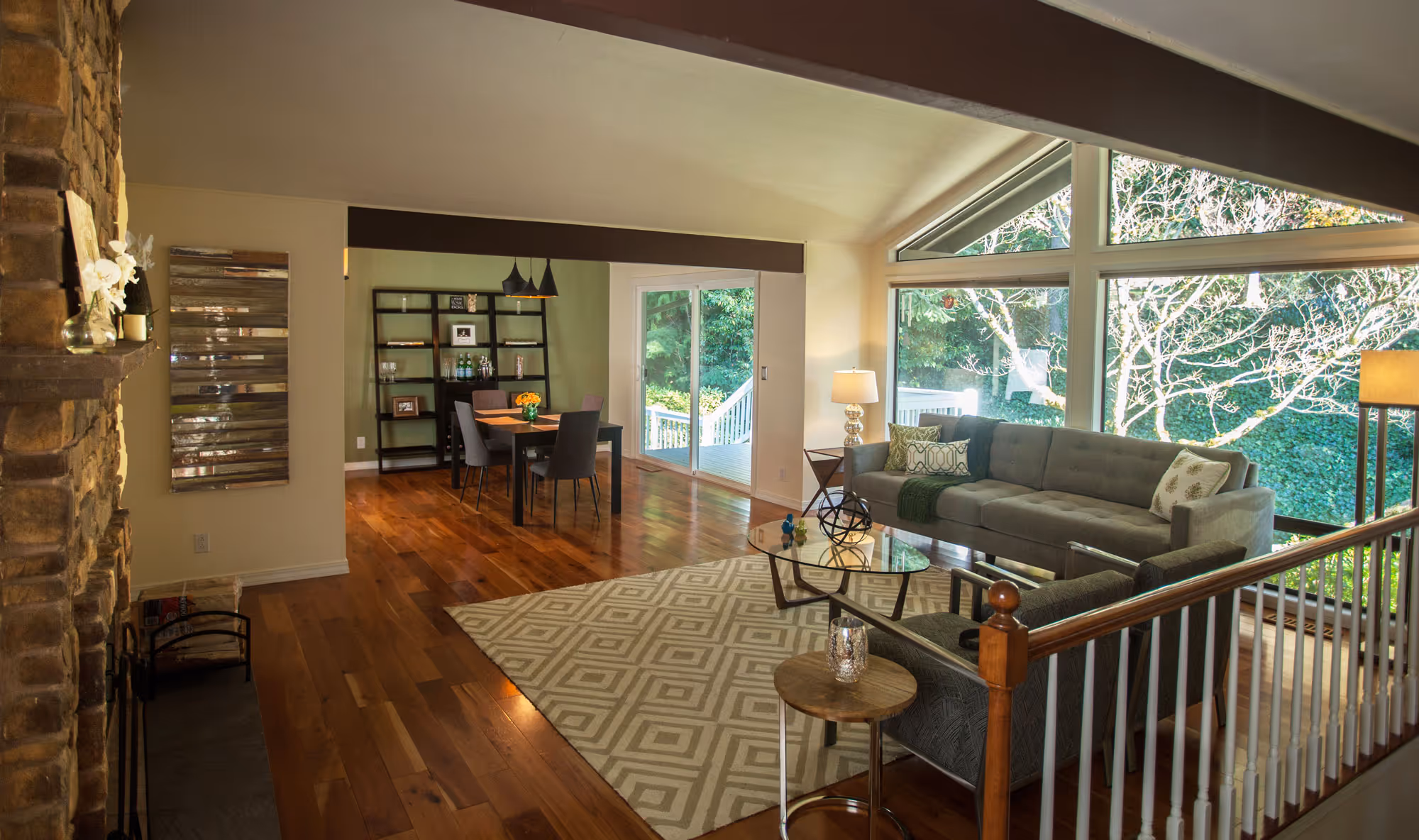 A cozy living and dining area with warm wood flooring and vaulted ceilings. The living room features a large gray sectional sofa with decorative pillows, a glass coffee table, and a floor lamp, all set on a patterned area rug. Large windows and a sliding glass door provide ample natural light and views of lush greenery outside. The dining area includes a dark wood table and chairs, with a shelving unit against the wall displaying various decor items. A stone fireplace is visible on the left, adding a rustic touch to the space. The overall atmosphere is inviting and comfortable.