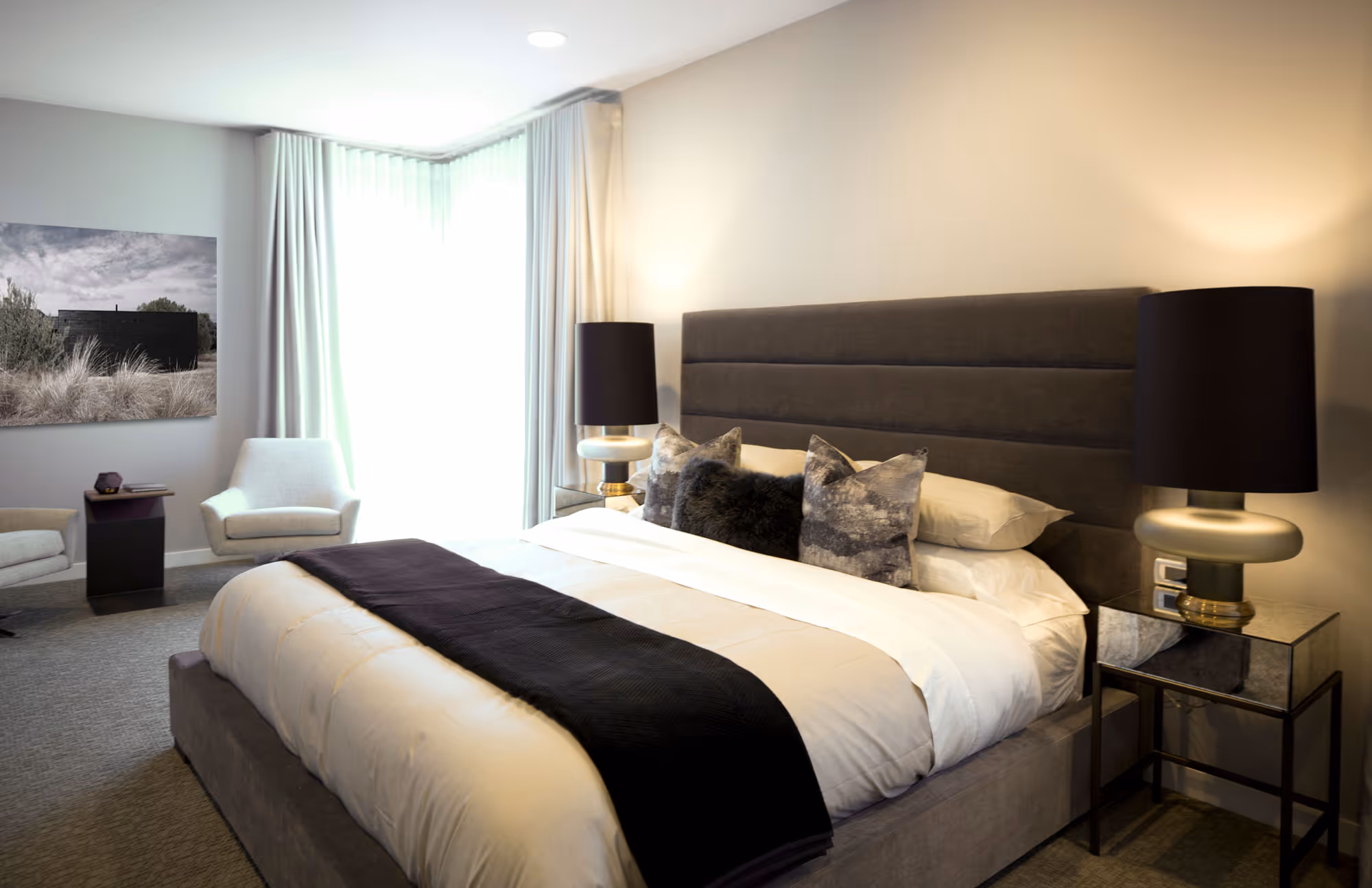A modern and elegant bedroom featuring a large bed with a padded gray headboard and white bedding, accented with dark gray and black pillows and a matching throw blanket. On either side of the bed are sleek bedside tables with black lamps featuring gold accents. The room has soft, neutral carpeting and light gray walls, with a large window covered by sheer white curtains that allow natural light to fill the space. In the corner, there are two white armchairs and a small side table, creating a cozy sitting area. A black and white photograph hangs on the wall, adding a touch of artistic flair to the room.