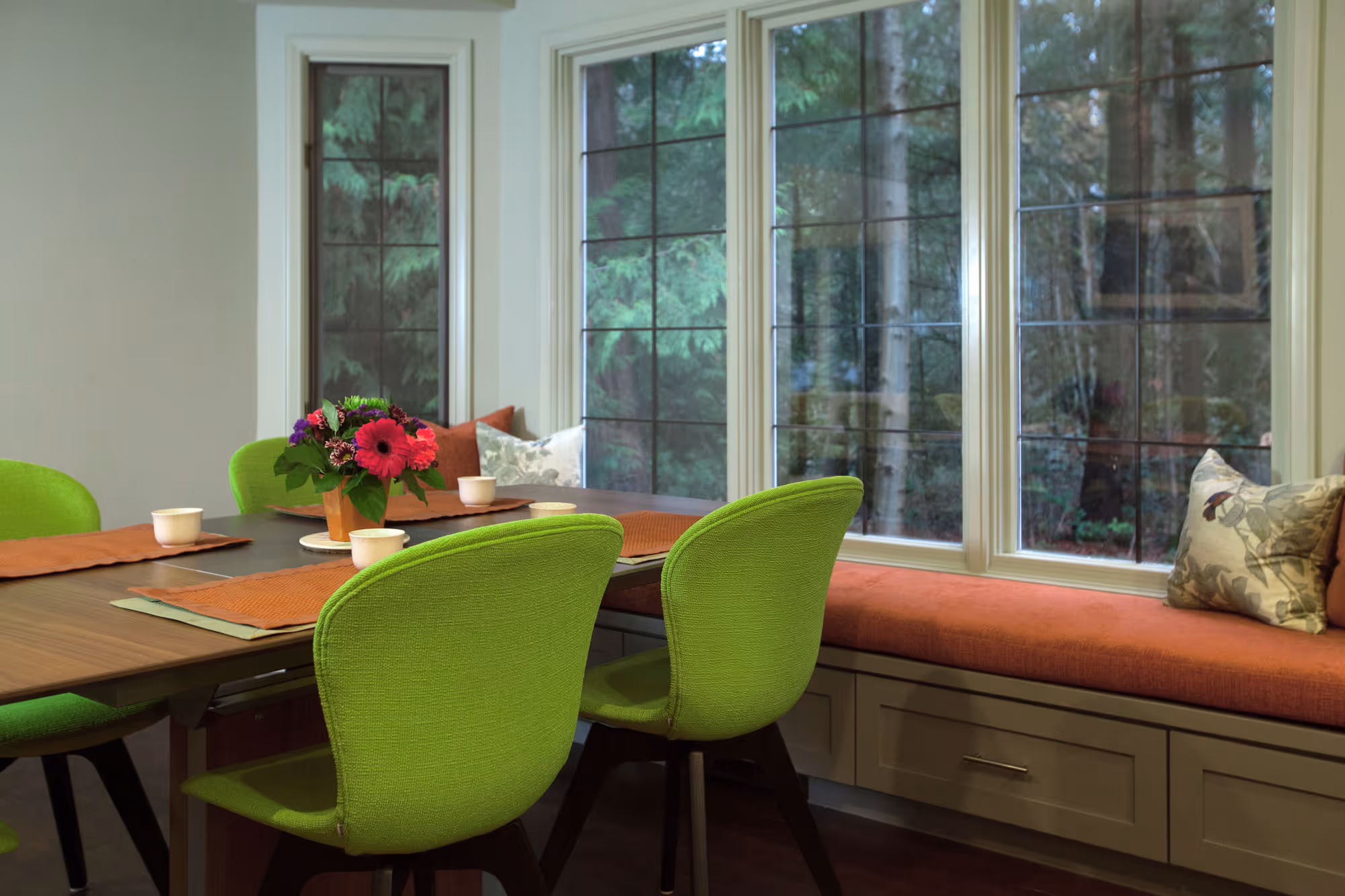 A cozy dining area featuring a wooden table with green upholstered chairs. The table is set with orange placemats and white cups, and a vase with vibrant flowers serves as the centerpiece. Adjacent to the table, a built-in bench with orange cushions and decorative pillows runs along a large set of windows, providing a view of the lush greenery outside. The room has a warm and inviting atmosphere, with natural light filtering through the windows and highlighting the green and orange color scheme.