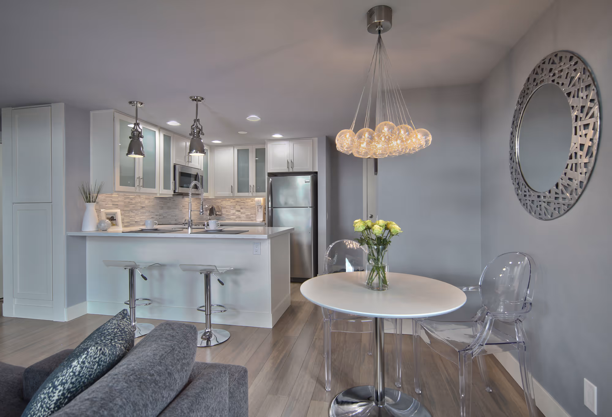 A modern, compact kitchen and dining area with a light and airy feel. The kitchen features white cabinetry, a stainless steel refrigerator, and a small island with a white countertop and bar stools. Pendant lights hang over the island, adding a stylish touch. The backsplash is made of light-colored tiles, complementing the sleek design. Adjacent to the kitchen is a small dining area with a round white table, clear acrylic chairs, and a decorative chandelier with multiple glass spheres. A large, decorative mirror hangs on the gray wall, and a small vase of yellow flowers sits on the dining table. The living area with a gray sofa is partially visible in the foreground.