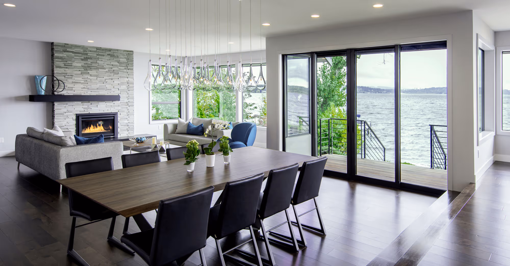 A modern open-concept living and dining area with a clean, contemporary design. The space features a comfortable gray sofa with blue accent pillows, a stylish blue swivel chair, and a sleek coffee table adorned with decorative candles. The dining area includes a wooden table with black chairs, illuminated by an elegant chandelier. The room is well-lit with recessed lighting, and the walls are decorated with abstract artwork. The hardwood floors and modern staircase add to the sophisticated ambiance of the home.