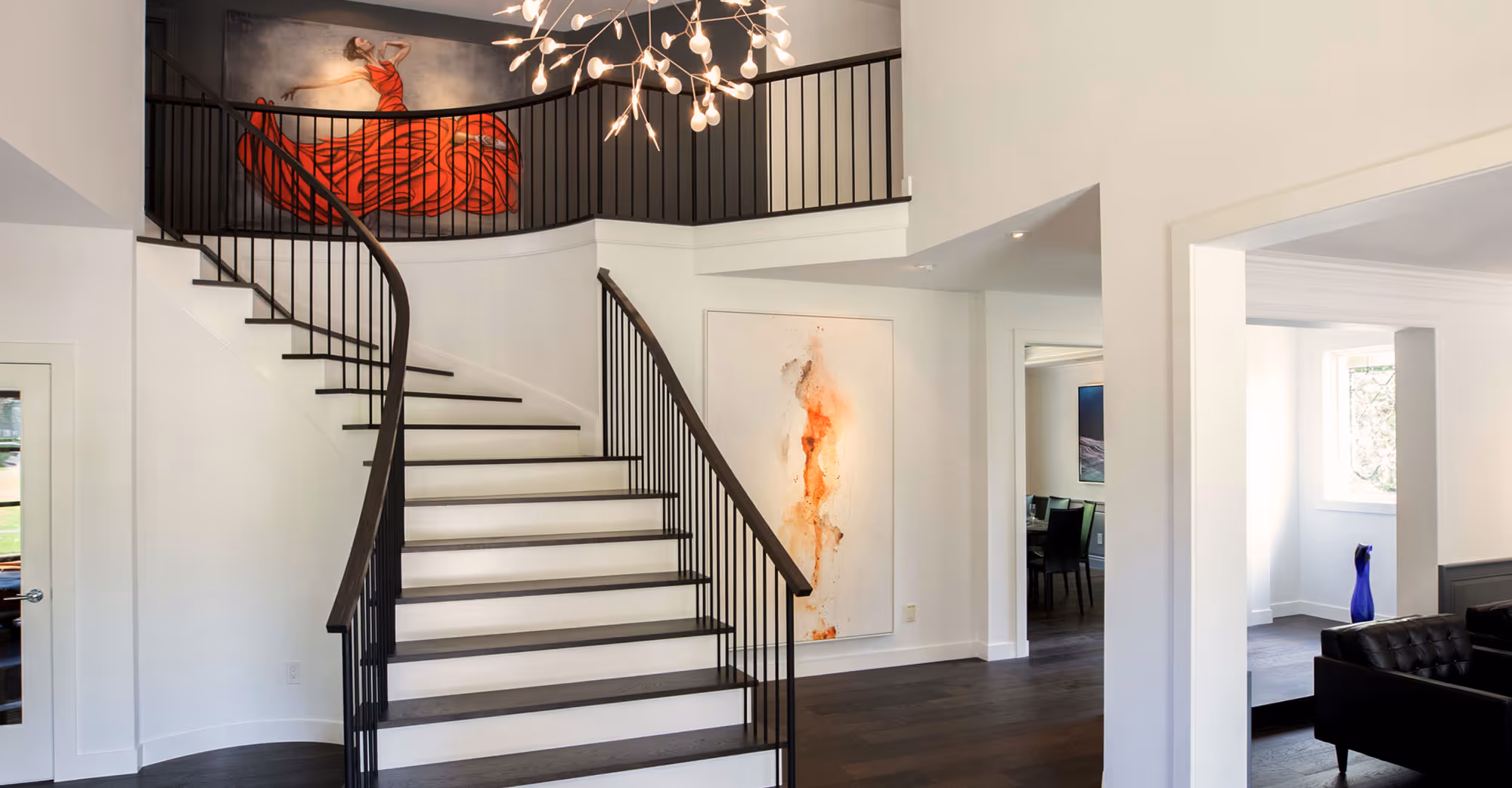 A stylish and modern foyer featuring a grand curved staircase with dark wood steps and a black railing. The space is highlighted by contemporary artwork, including a striking painting of a woman in a red dress above the landing and an abstract piece with orange and brown tones on the wall. The foyer is bright with white walls and dark wood flooring, creating a sharp contrast. A unique chandelier with multiple small bulbs hangs from the ceiling, adding an artistic touch. Adjacent rooms, including a dining area and a sitting room with black leather chairs, are visible through open doorways.