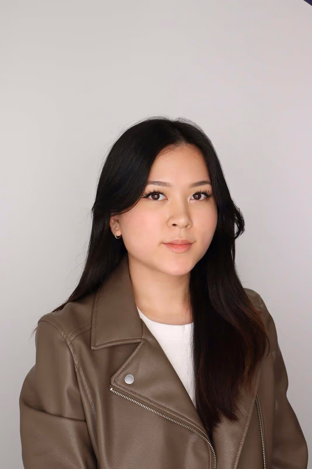 Portrait of a young woman with long black hair wearing a brown leather jacket and white top against a plain background.