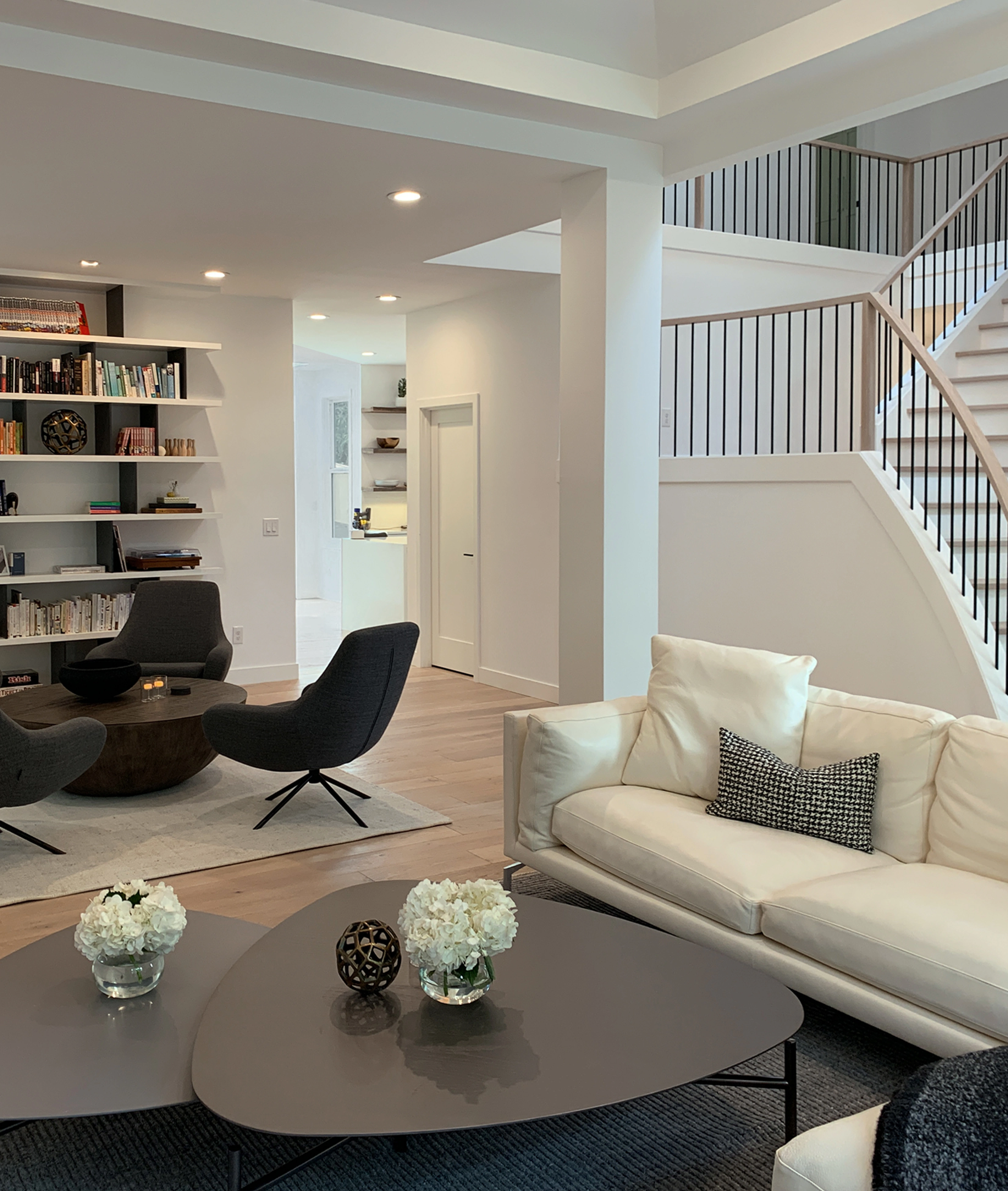 Modern living room with a gas fireplace, wall-mounted TV, dark coffee tables with white flower vases, gray armchairs, and a bookshelf filled with books.