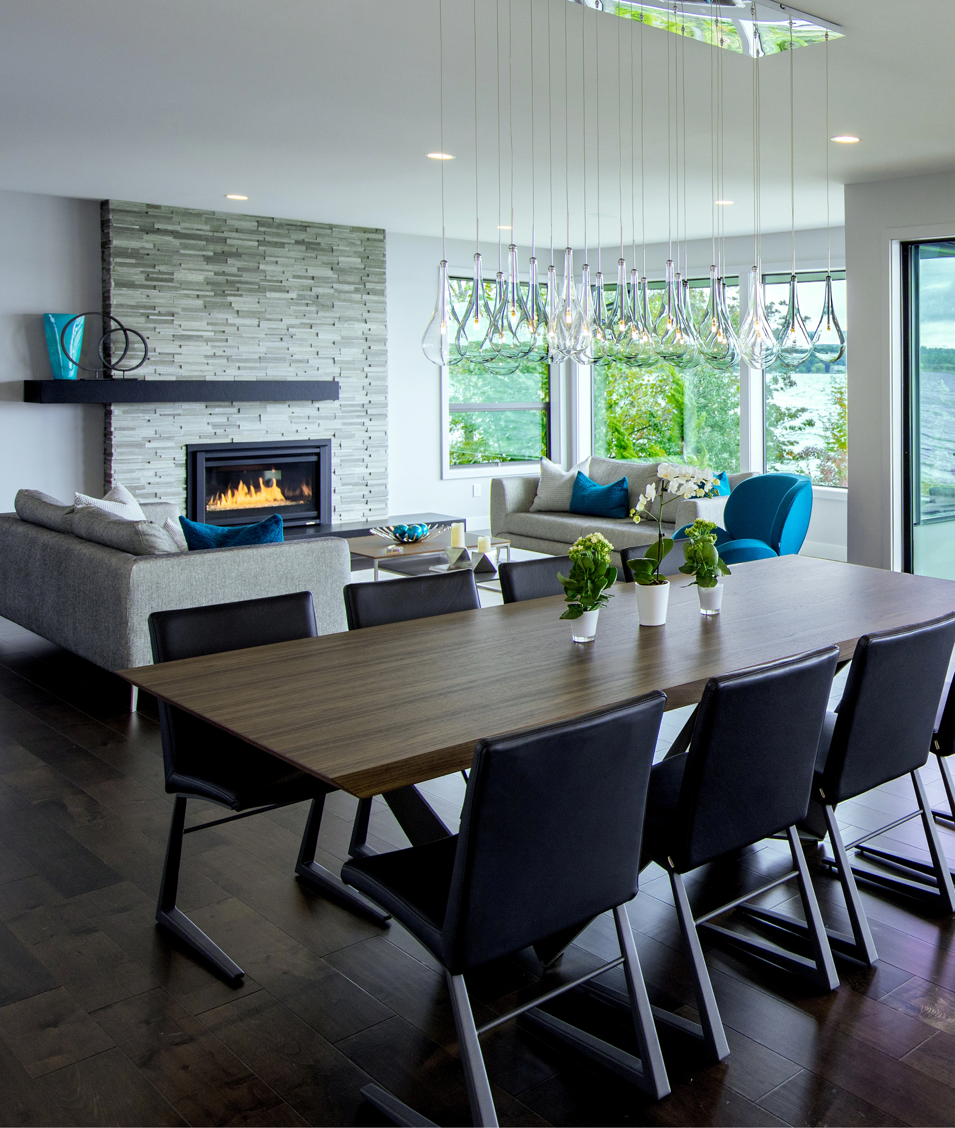 Modern dining and living room with wooden table, black chairs, hanging glass pendant lights, fireplace, and large windows overlooking waterfront.