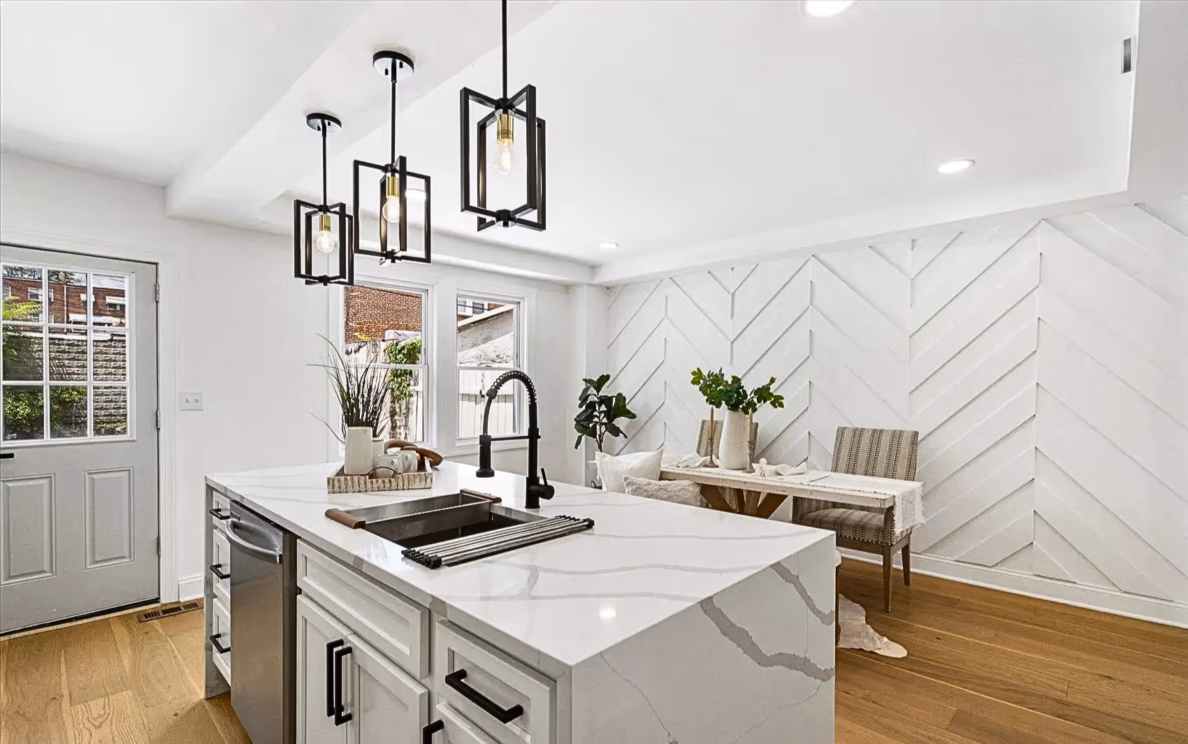 Stylish kitchen with white quartz countertops featuring grey veins, black sink faucet, and white cabinets with black handles. Three black lantern pendant lights hang above the kitchen island. The space opens to a dining area with a white accent wall showcasing a chevron wood plank pattern. Fresh windows and continuous plank wood flooring enhance the bright, cohesive design.