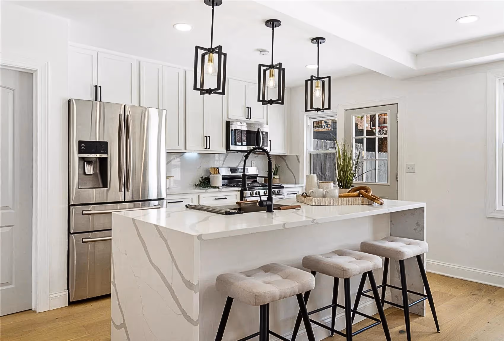 Contemporary kitchen featuring a large white quartz island with grey veins, complemented by a black sink faucet and white cabinets with black handles. Three black lantern pendant lights hang above the island. Stainless steel appliances, newly installed grey back door, and plank wood flooring throughout complete the modern design.