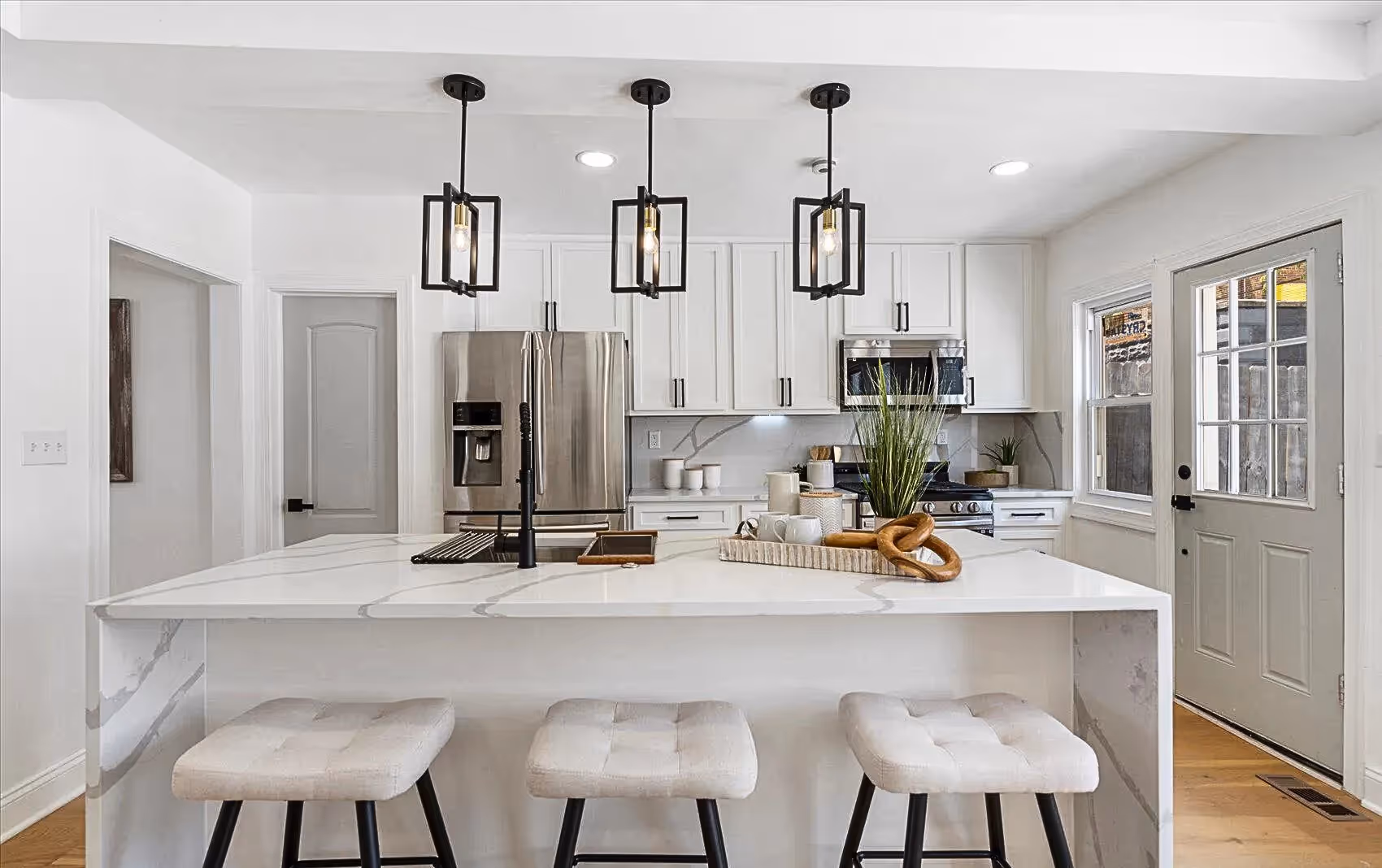 Modern kitchen featuring a large white quartz countertop with three black pendant lights hanging above. Stainless steel appliances, recessed ceiling lighting, and an open layout leading to the living area.