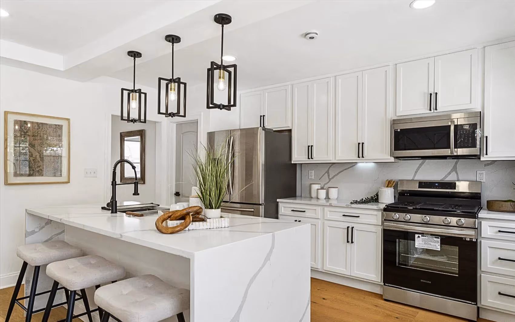 Spacious kitchen showcasing a sleek white quartz island beneath three modern black pendant lights. Features include stainless steel appliances, recessed lighting, and a seamless transition to the adjacent living space.
