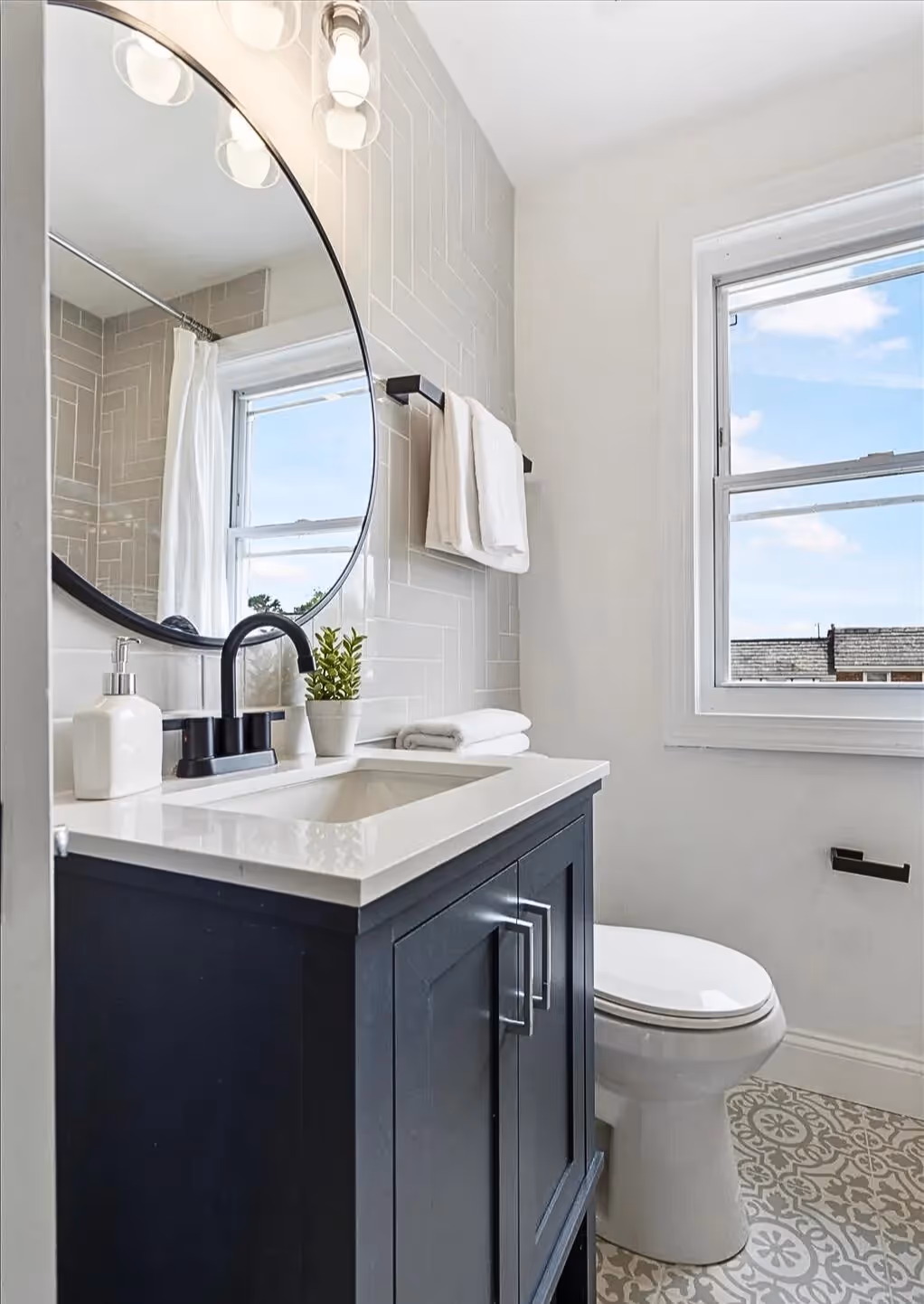 Bathroom featuring a deep blue vanity with a black circular mirror and vanity lights above. White walls and a new window complete the clean, modern design