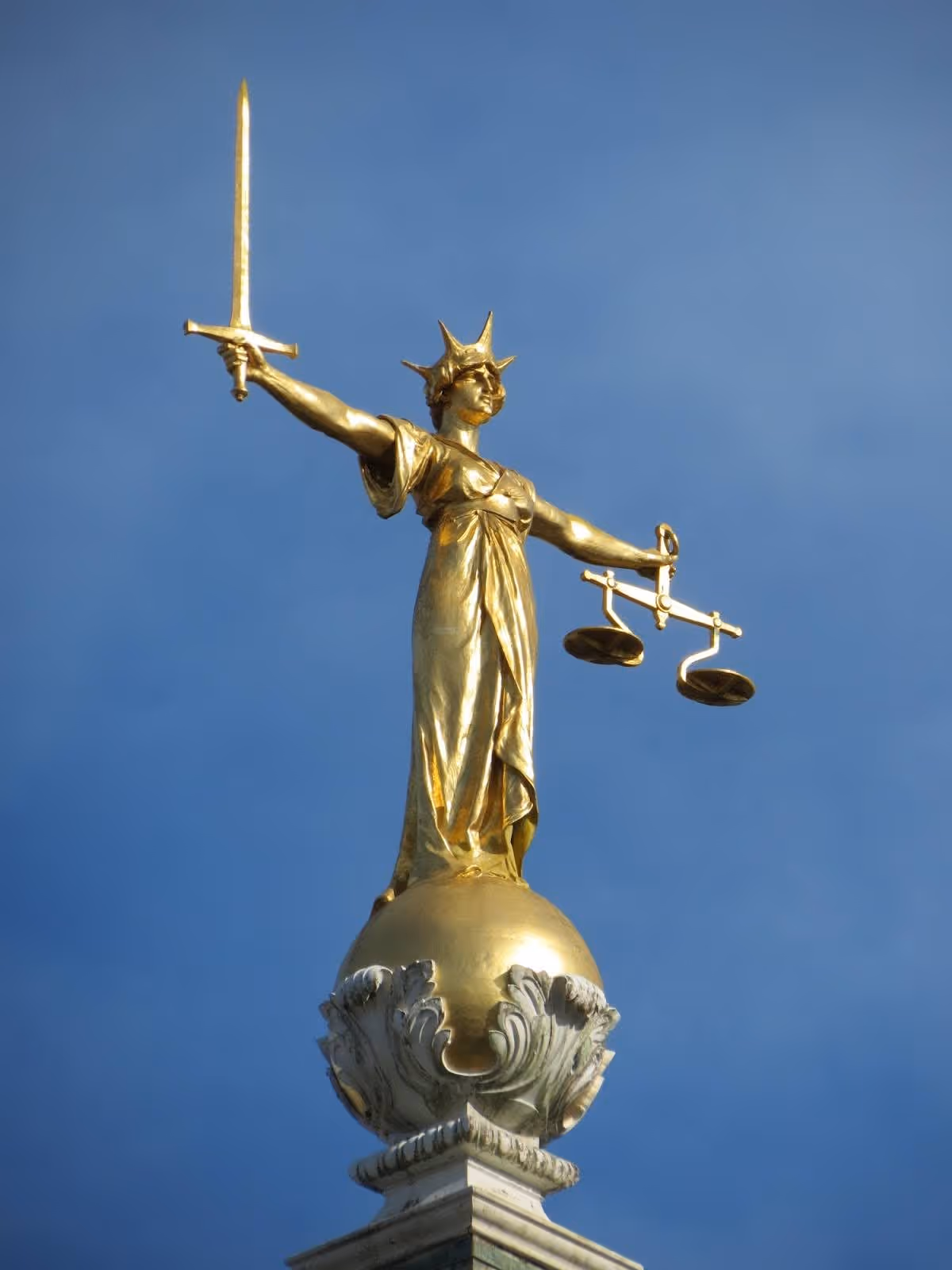 A golden statue of Lady Justice holding a sword and scales stands atop a dome against a clear blue sky, representing fairness and law.