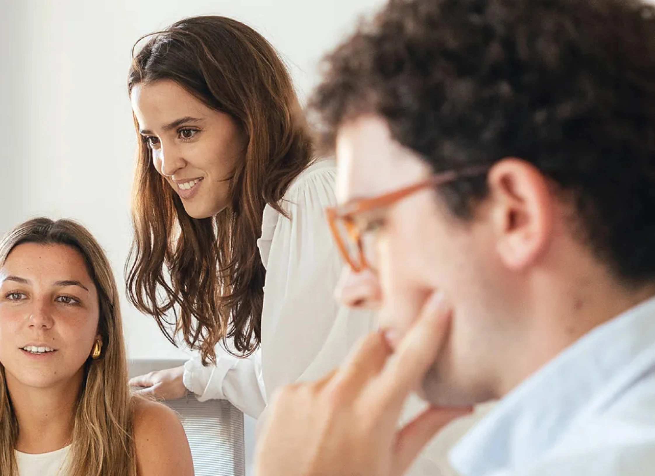 Tres personas jóvenes en una reunión de trabajo, conversando y colaborando en una oficina luminosa.