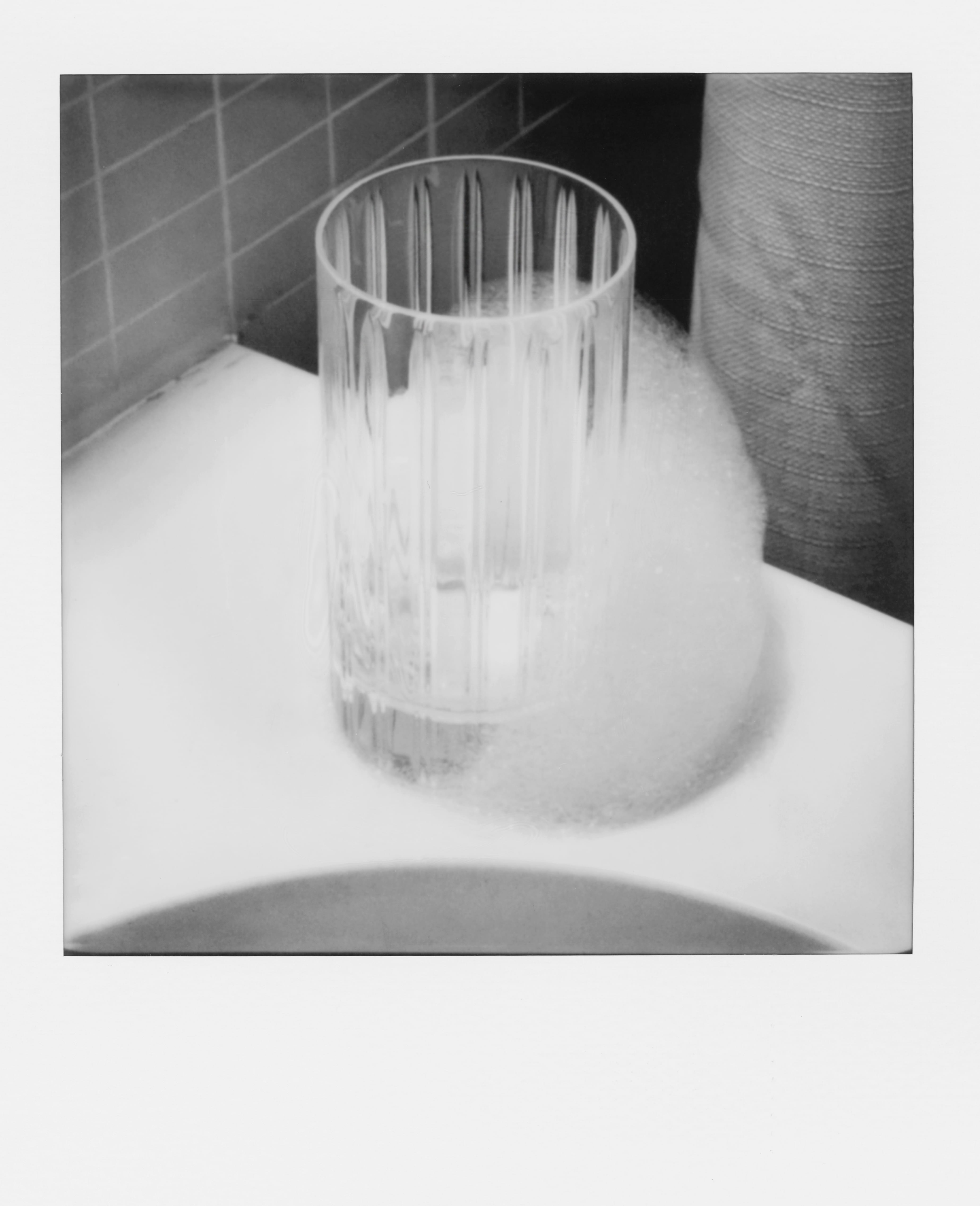 Clear glass on a white surface surrounded by soap foam near tiled wall and fabric.