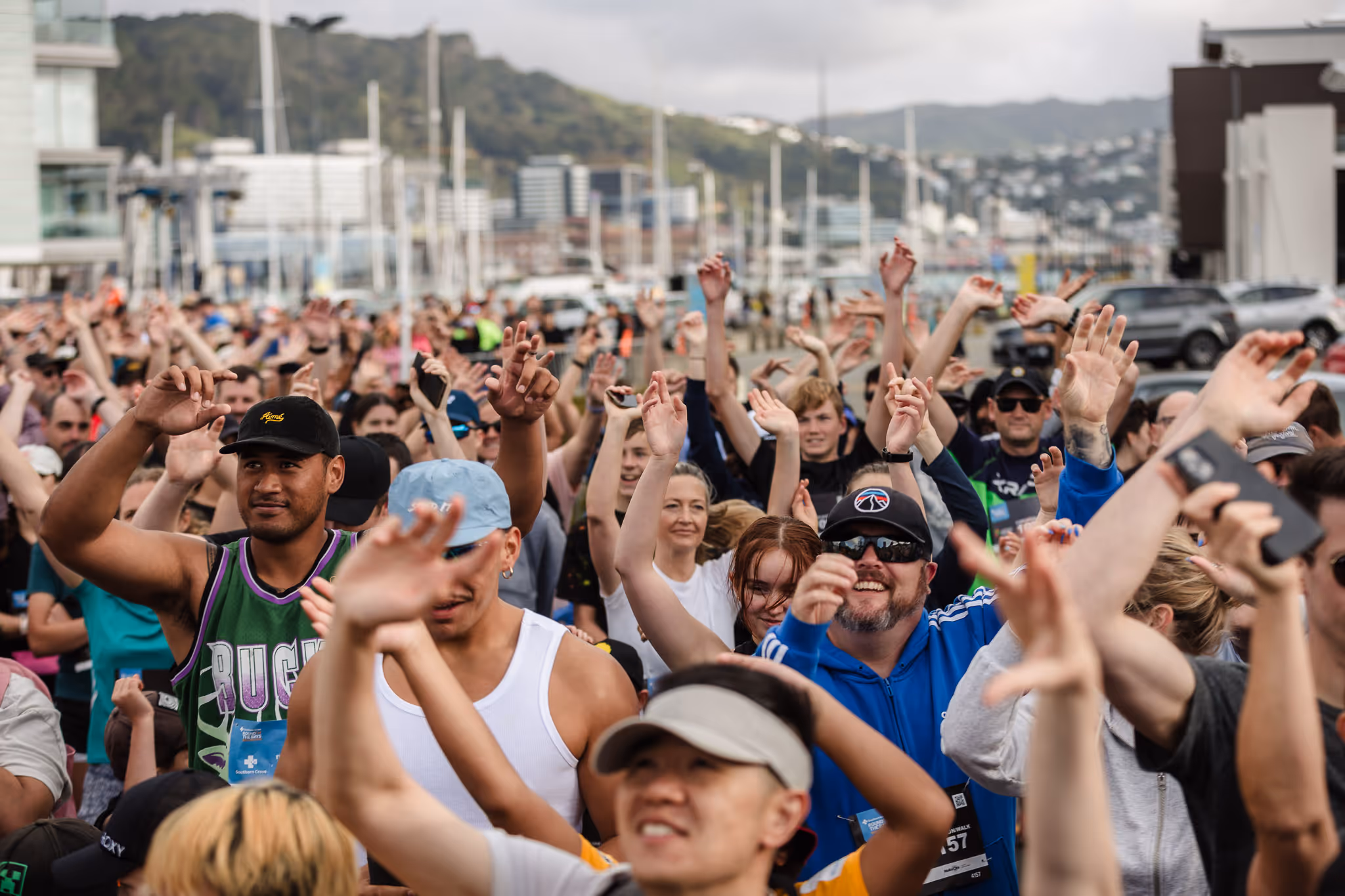 A large crowd of enthusiastic people with raised hands gather outdoors on a cloudy day. The scene is vibrant, with individuals of various ages, many wearing casual and sports attire, suggesting a lively public event or gathering. Urban buildings are visible in the background.
