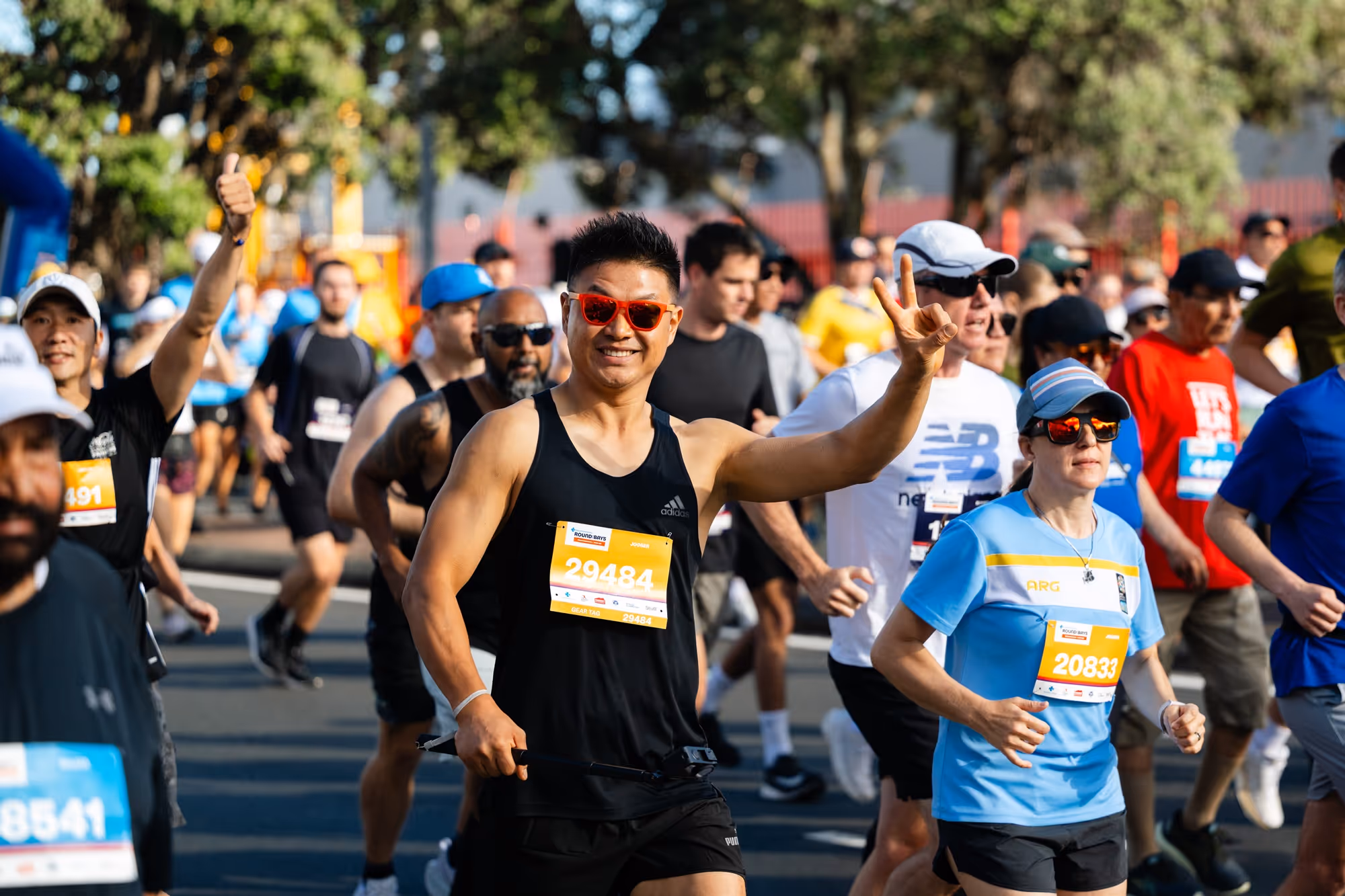 ‍A large group of runners is captured at the start line on a bustling city street in Auckland New Zealand. Men women children wheelchair users are participating, with some wearing race bibs and athletic gear. The background shows a mix of scenery and age groups, with a clear, sunny sky overhead.