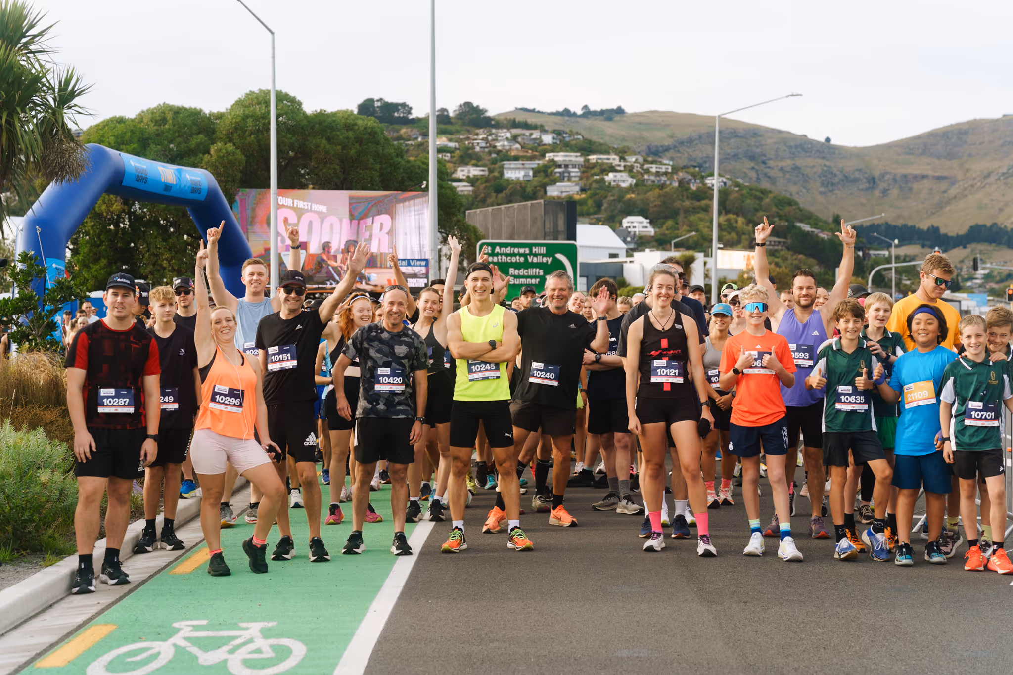 A large group of runners is captured mid-race on a bustling city street in Christchurch New Zealand. Both men and women are participating, with some wearing race bibs and athletic gear. The background shows a mix of scenery and age groups, with a clear, sunny sky overhead.