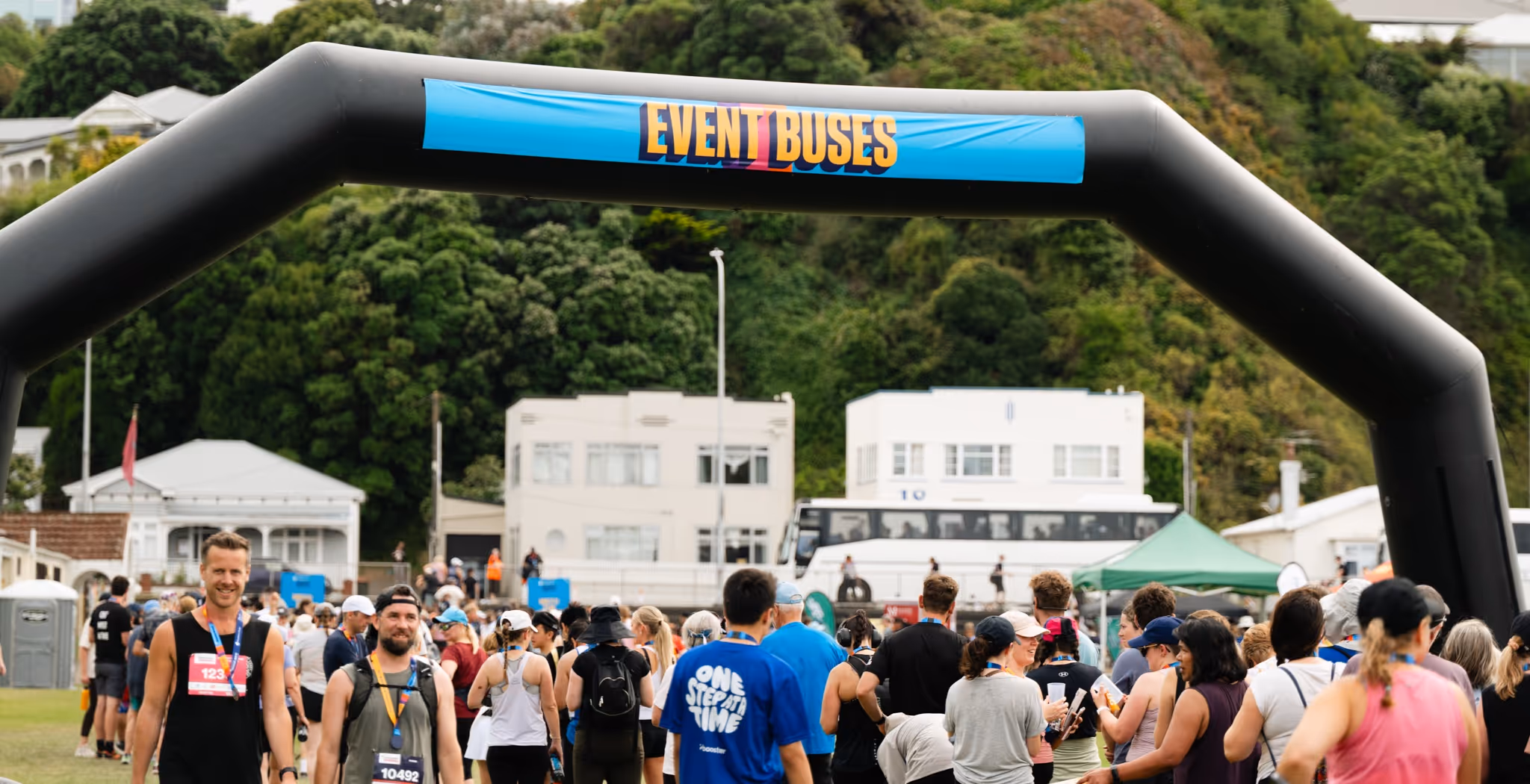 A group of runners gather at the starting line of a race. The large yellow inflatable archway reads "START" and "Nuku Ora." The background shows a waterfront area with boats, buildings, and a cloudy sky.