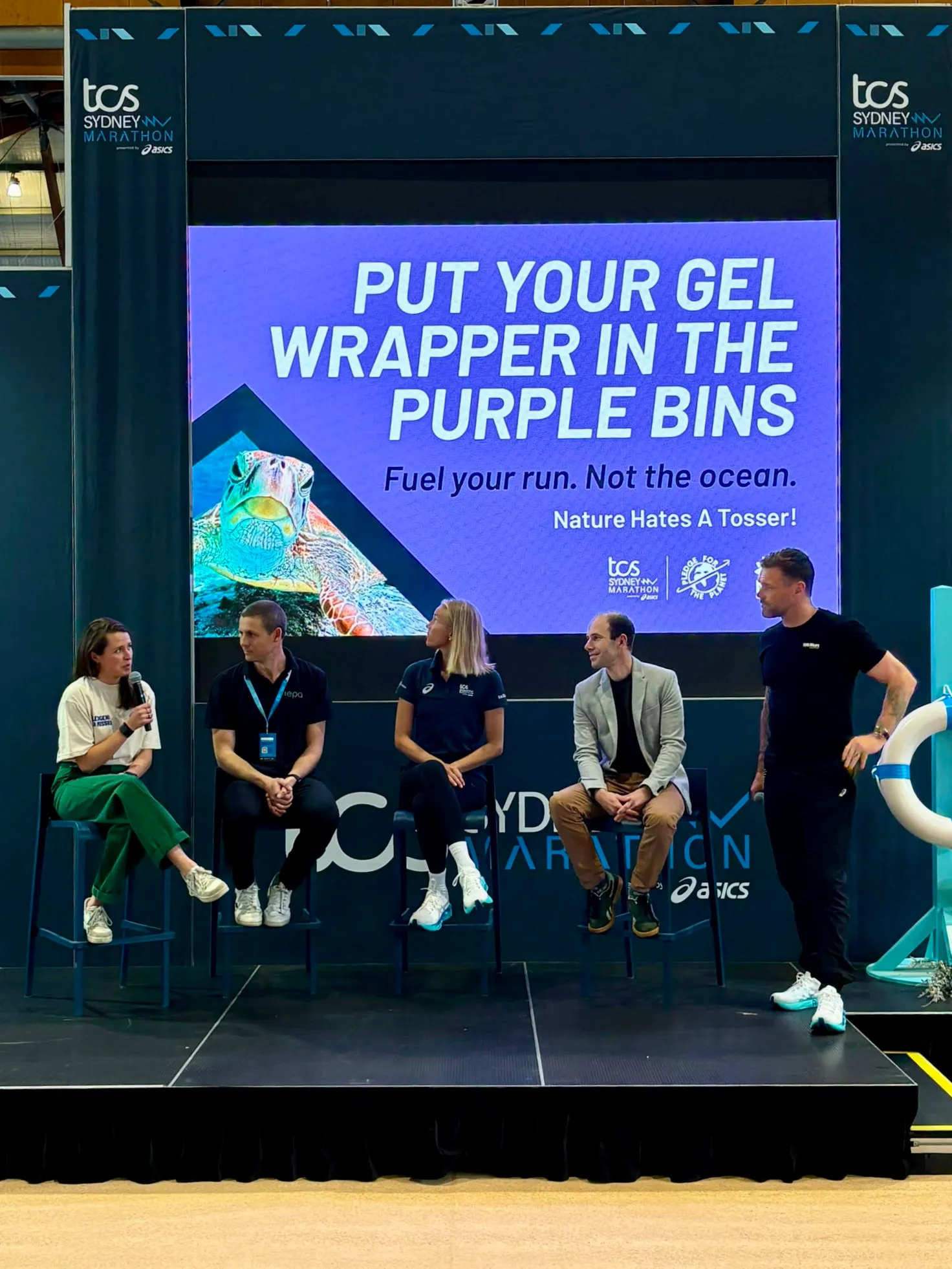 Panel on stage at the TCS Sydney Marathon expo under a screen reading 'Put your gel wrapper in the purple bins'