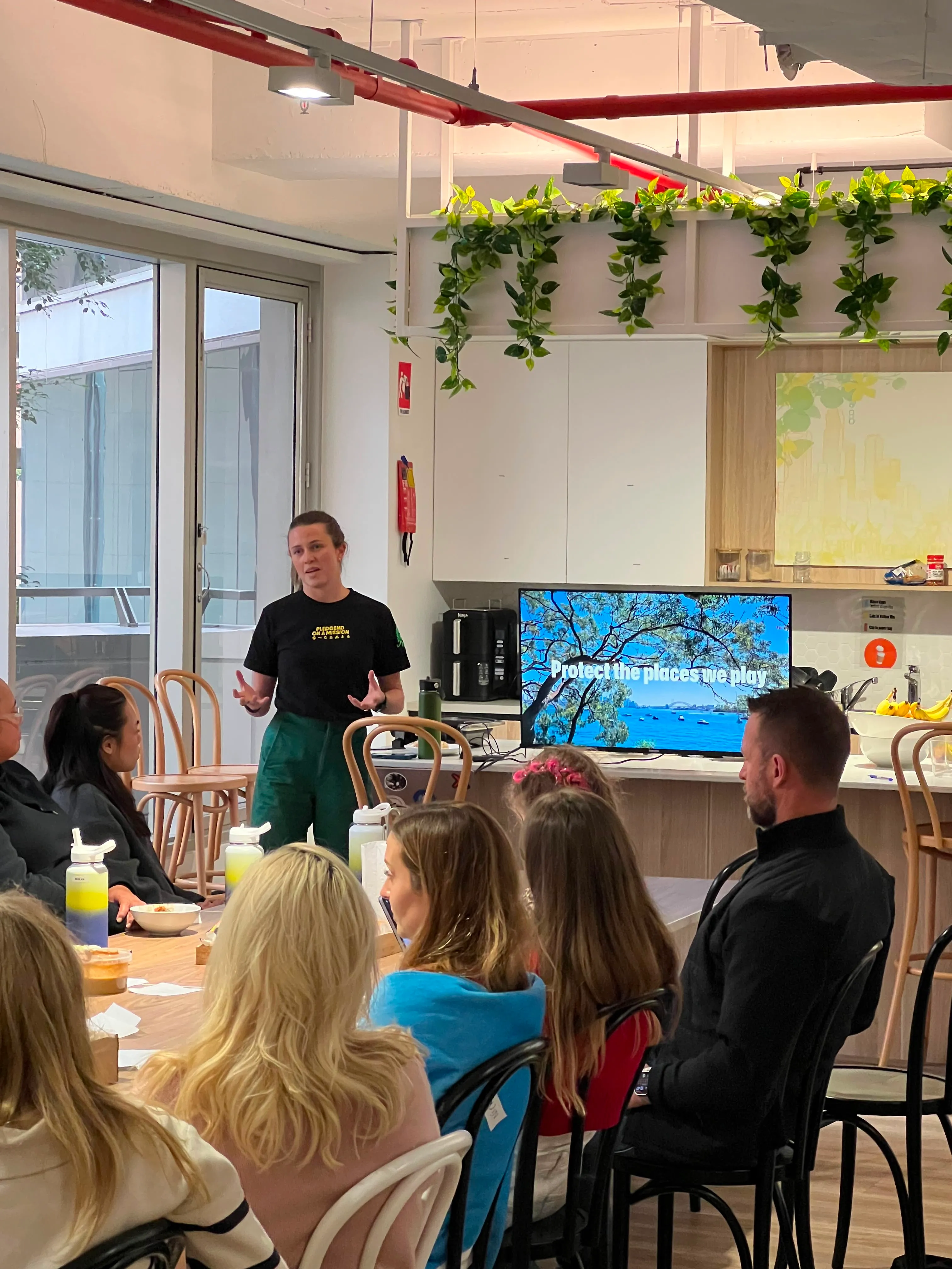 Speaker presenting to seated colleagues in an office kitchen beside a screen