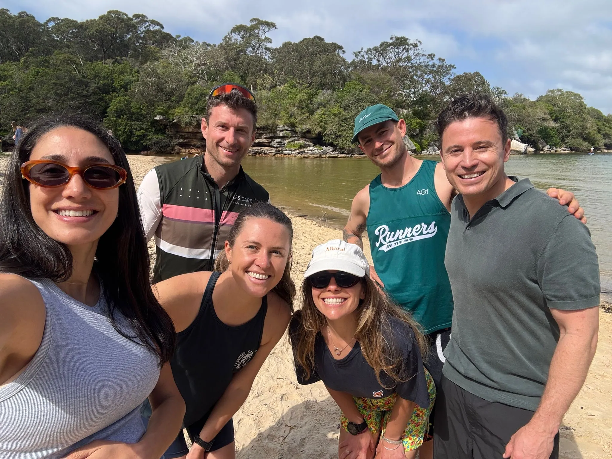 Five friends in activewear posing beside a bush-lined river beach