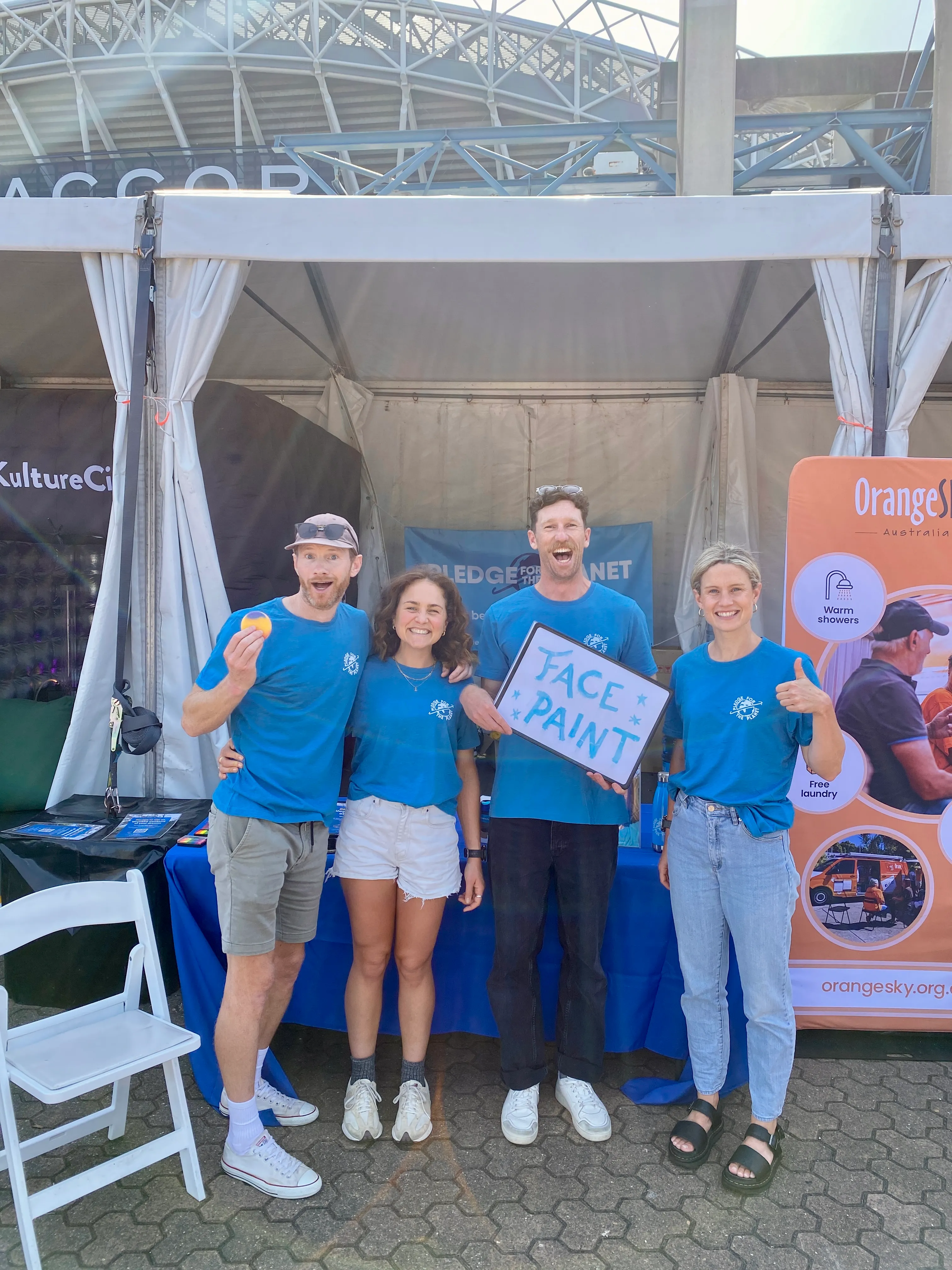 Four volunteers in matching tees at an event marquee holding a face paint sign