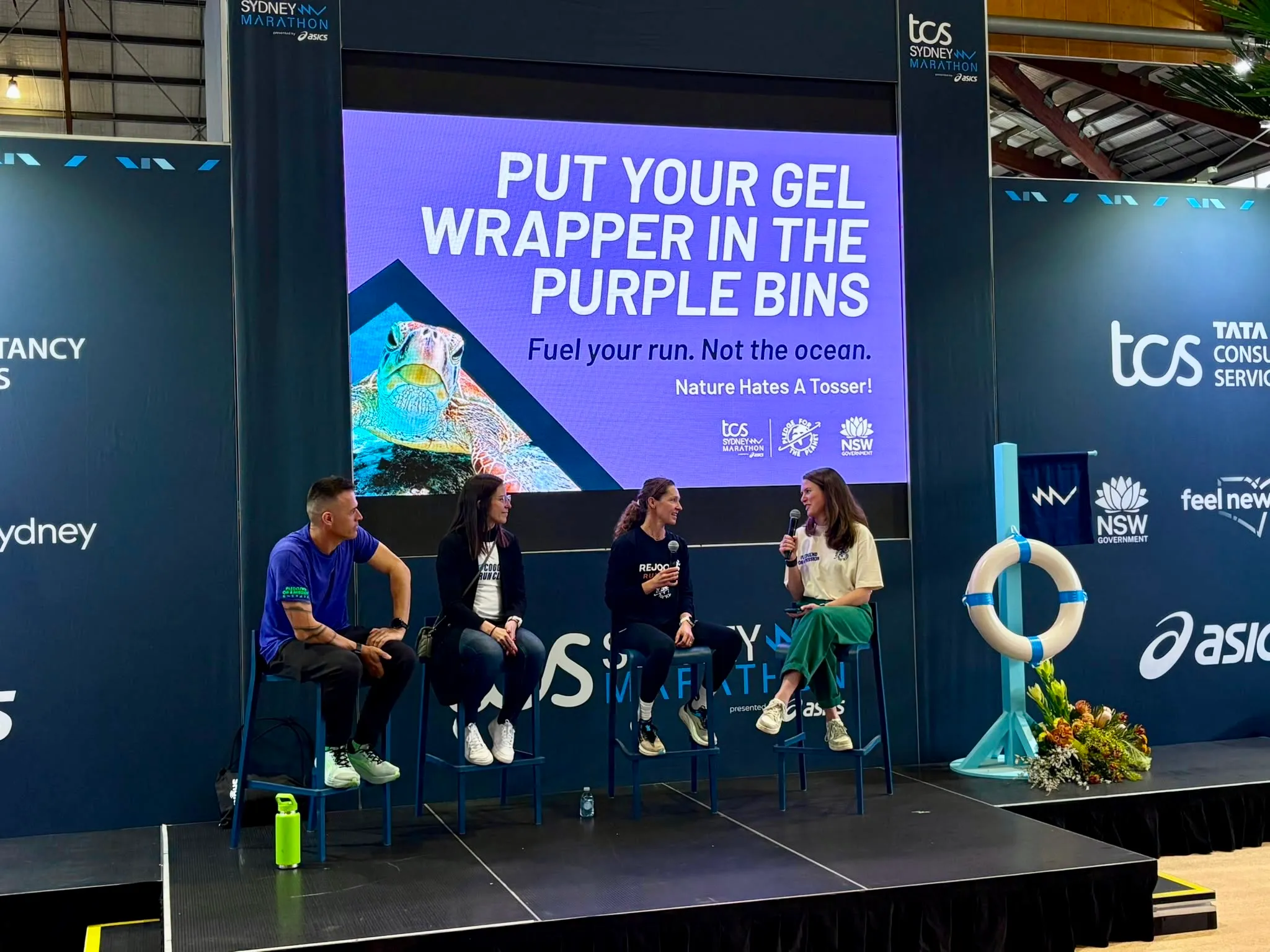 Four panellists on stage at a race expo beside a gel wrapper recycling message