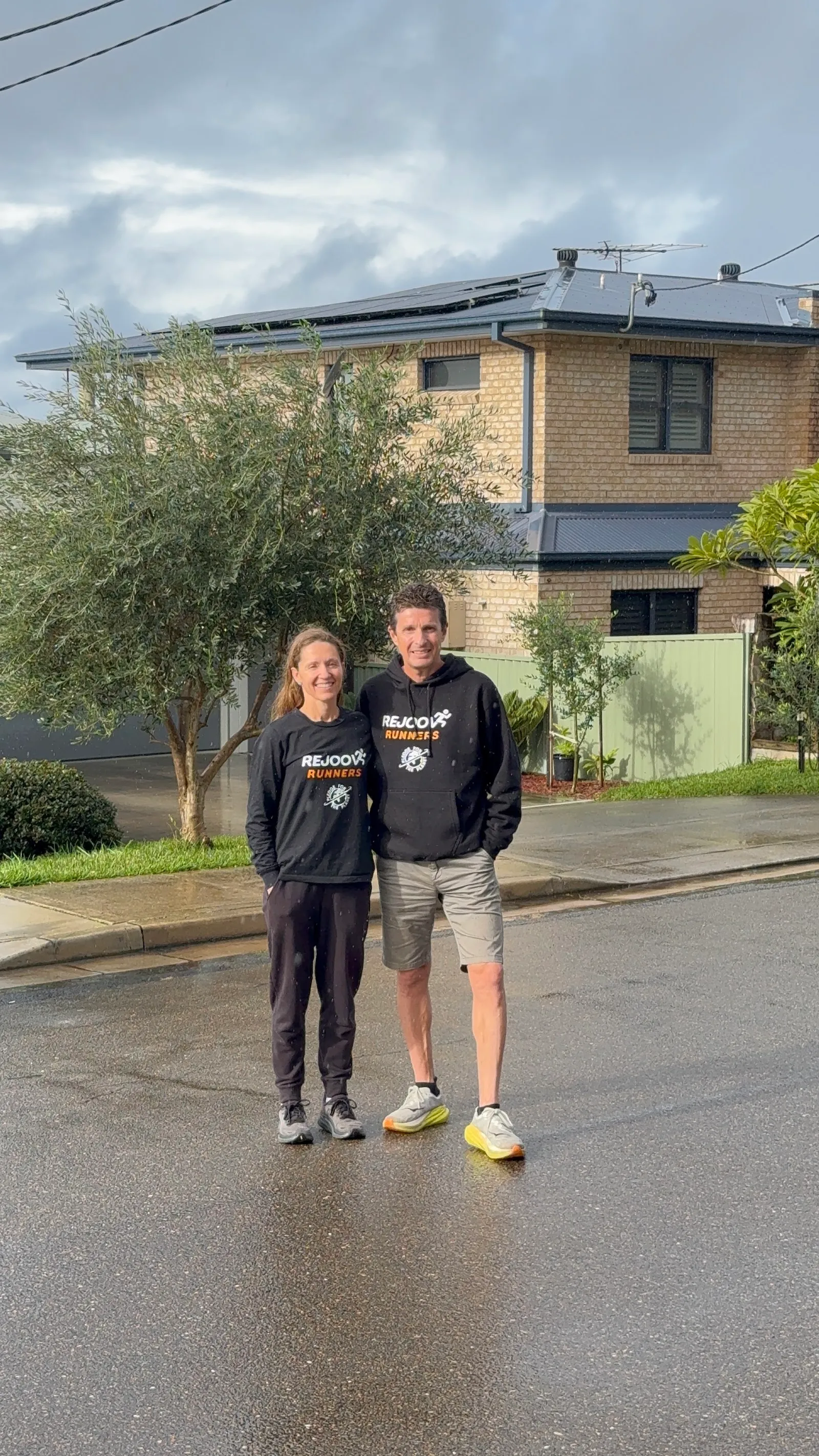 Greta and Chris Truscott outside their home in Rejoov Runners hoodies, with rooftop solar panels behind them