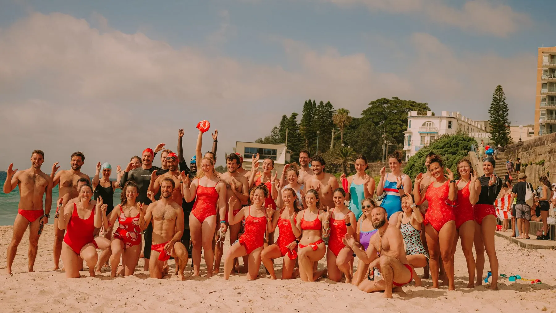 The Crusties ocean swimmers, many in matching red swimsuits, cheer together on the sand