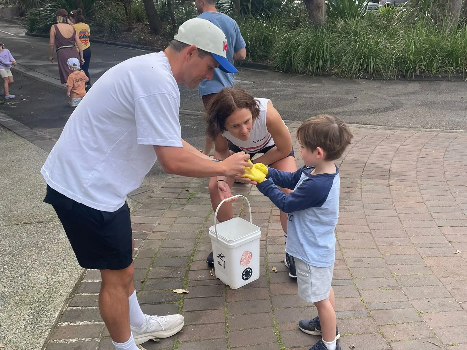 A young boy hands collected rubbish to two adults beside a bucket during a clean-up