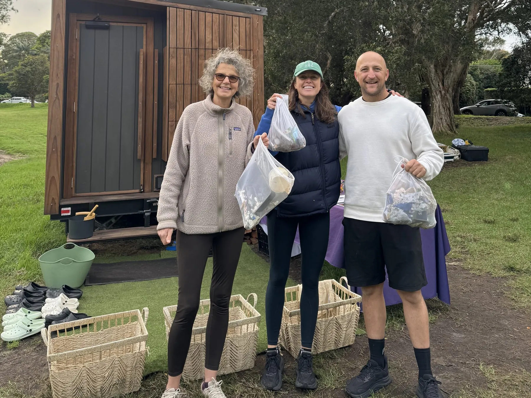 Three ploggers hold up bags of collected litter in front of The Sauna Project's timber mobile sauna