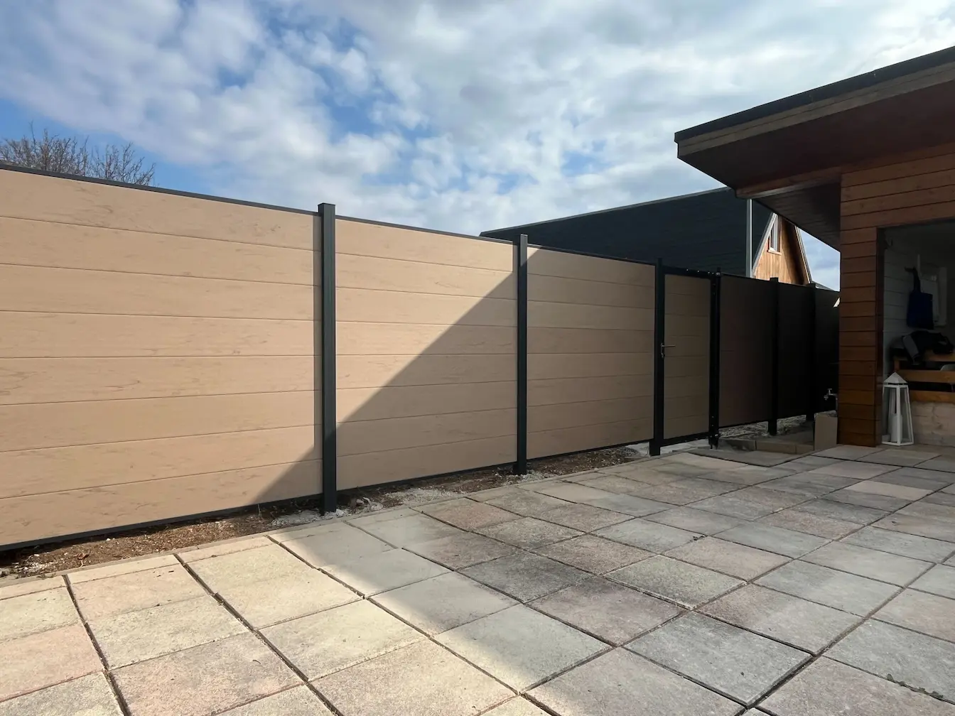 Moderner WPC-Sichtschutzzaun in Holzoptik/Beige mit anthrazitfarbenen Pfosten und einer passenden Gartentür neben einer gepflasterten Terrasse