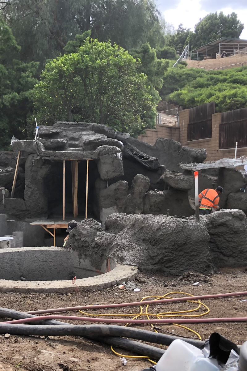 Workers are constructing a cave-like structure using cement, with trees visible in the background.