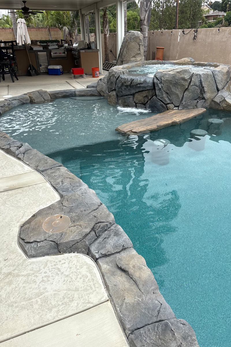 A swimming pool and an integrated jacuzzi, both constructed from stone. In the background, there's an outdoor kitchen in the patio area.