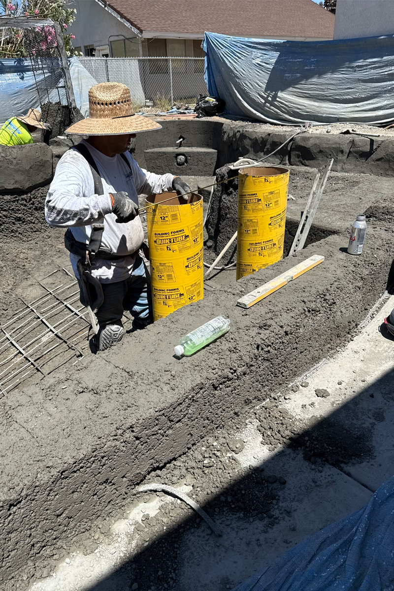 A worker is holding two yellow pipes inside a swimming pool that is currently under construction.