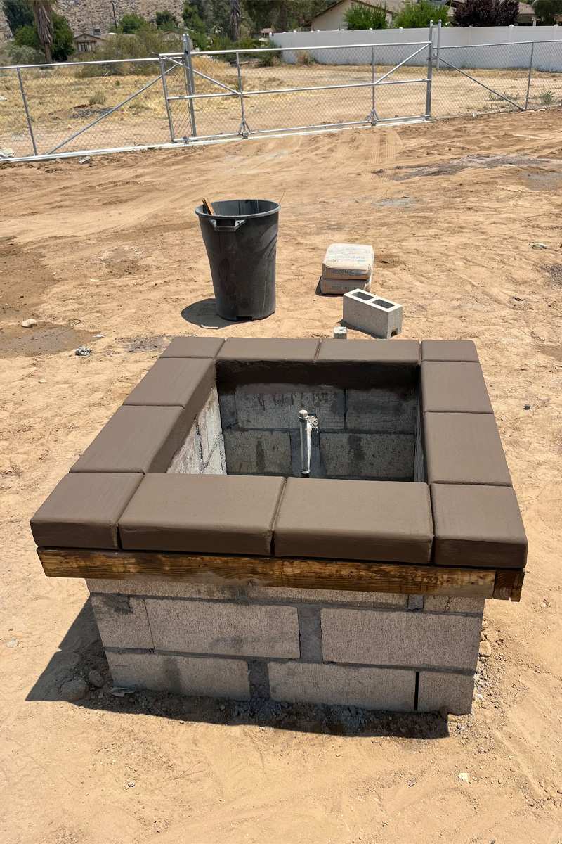 A fireplace currently under construction, surrounded by a dirt patio, with a metal fence in the background.