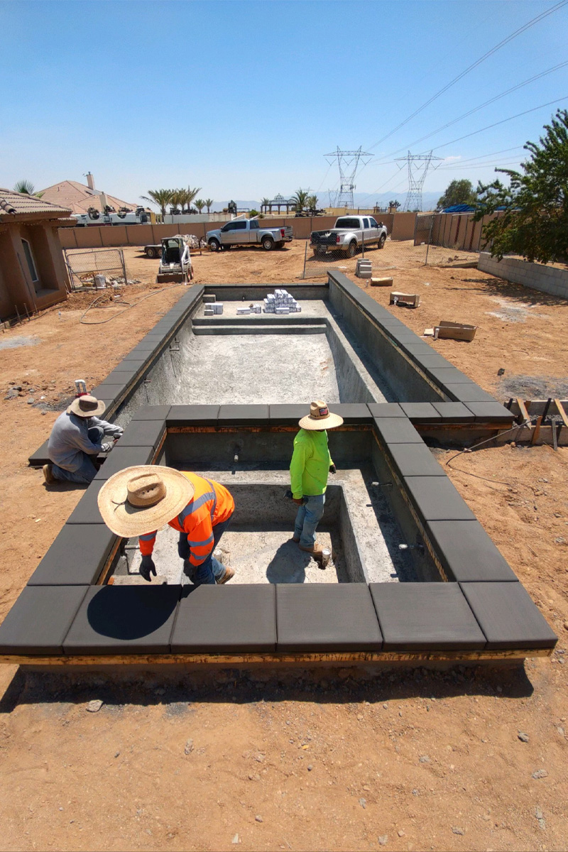 Three men are working on the construction of a swimming pool and an integrated jacuzzi. The area around them is a dirt patio, and pickup trucks are visible in the background.