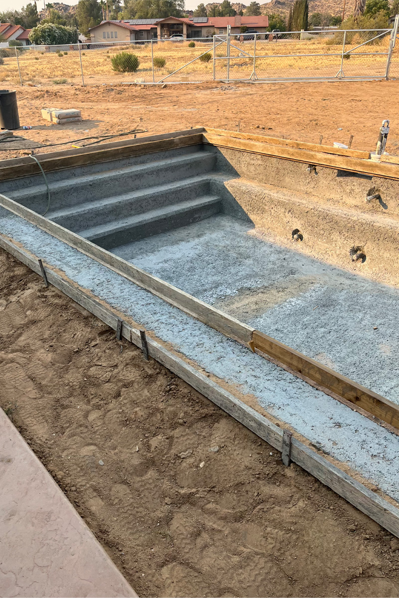A swimming pool currently under construction, with visible cement work, set within a vast, earthy desert area.