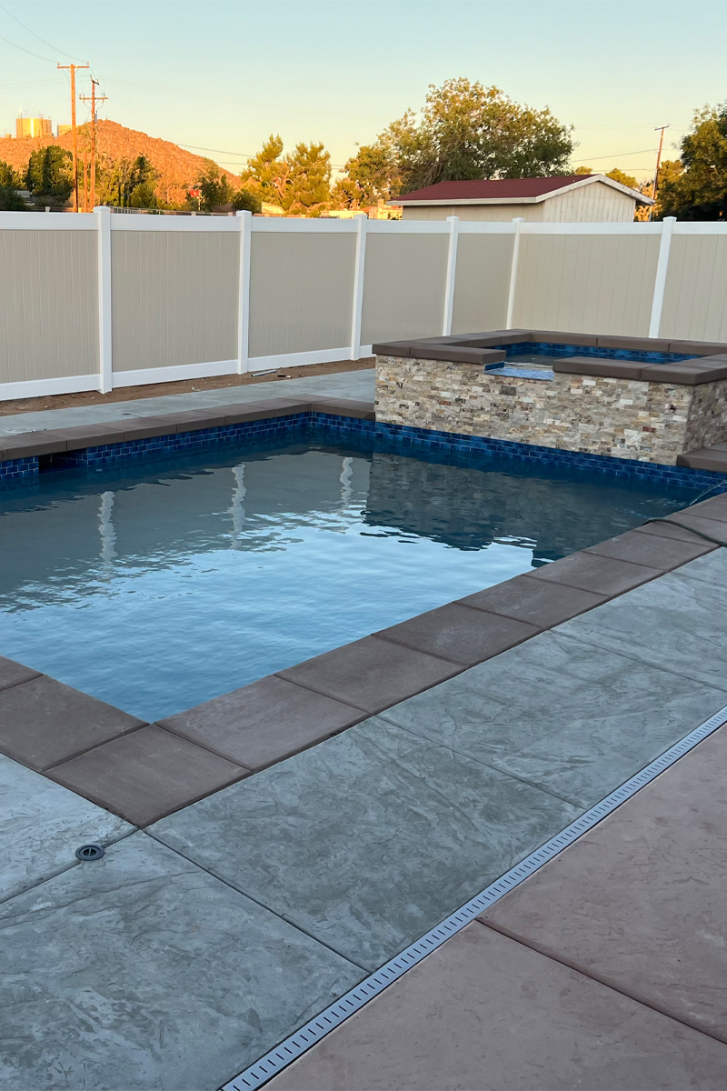 A square swimming pool with an integrated jacuzzi. In the background, there is a fence with a beige and white color scheme.