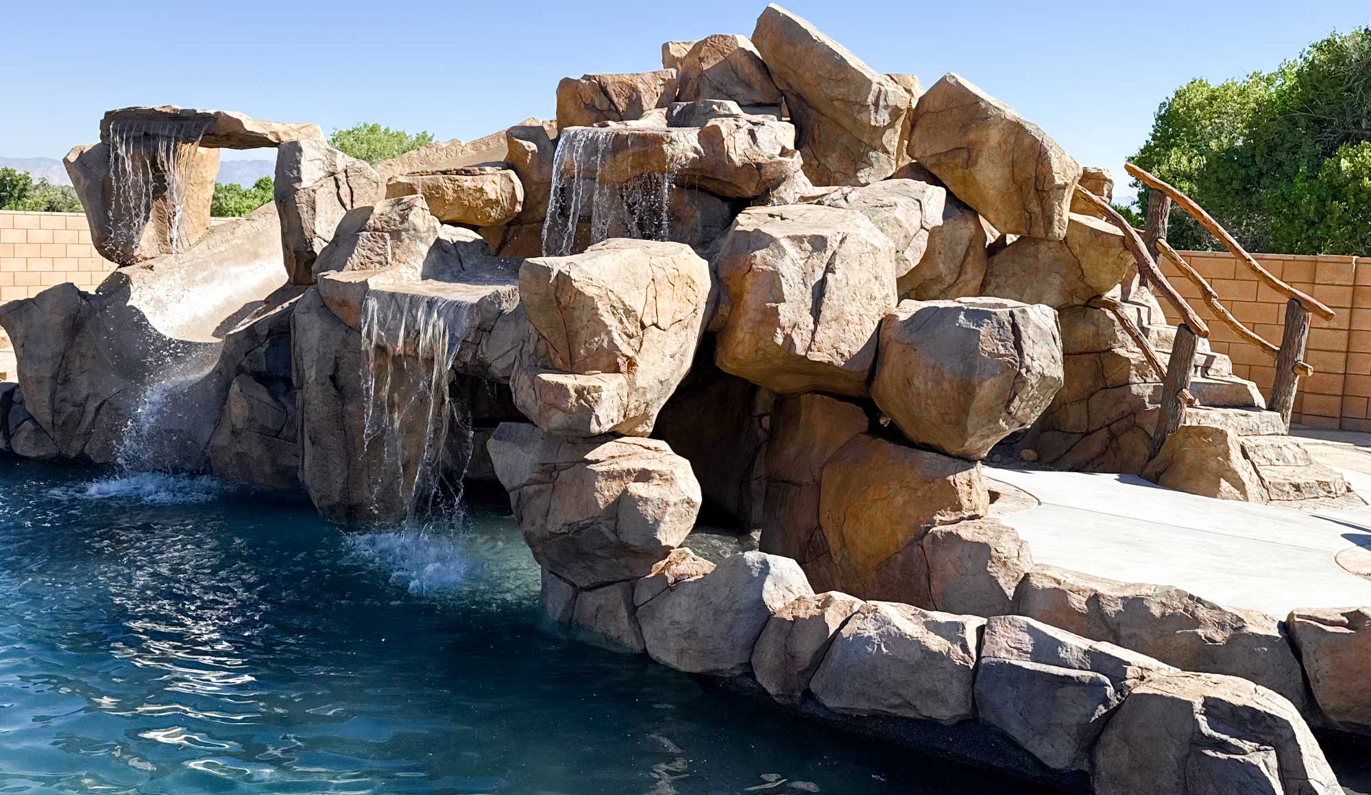 Three majestic and imposing waterfalls, constructed from natural stone, cascading into a swimming pool.

