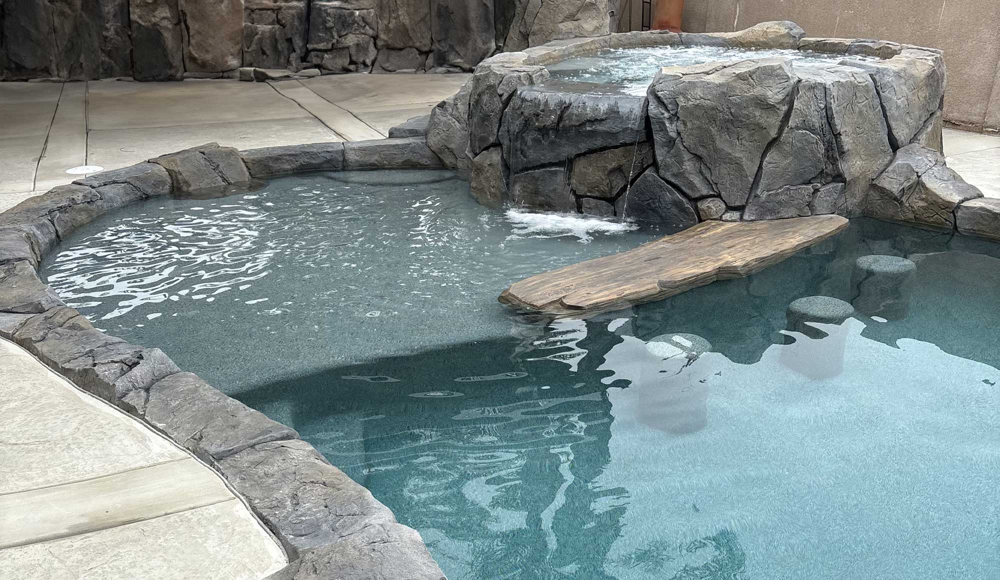 A swimming pool with an integrated jacuzzi from which a small waterfall descends. Both the jacuzzi and the waterfall are crafted from stones, complemented by a decorative wooden element.