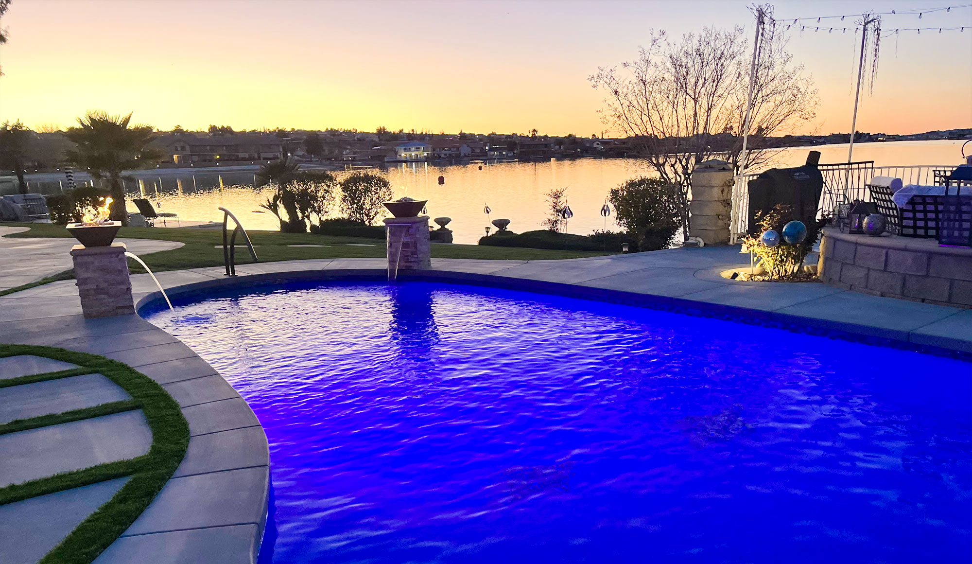 A beautiful blue swimming pool offering a stunning view of a lake and a picturesque sunset.

